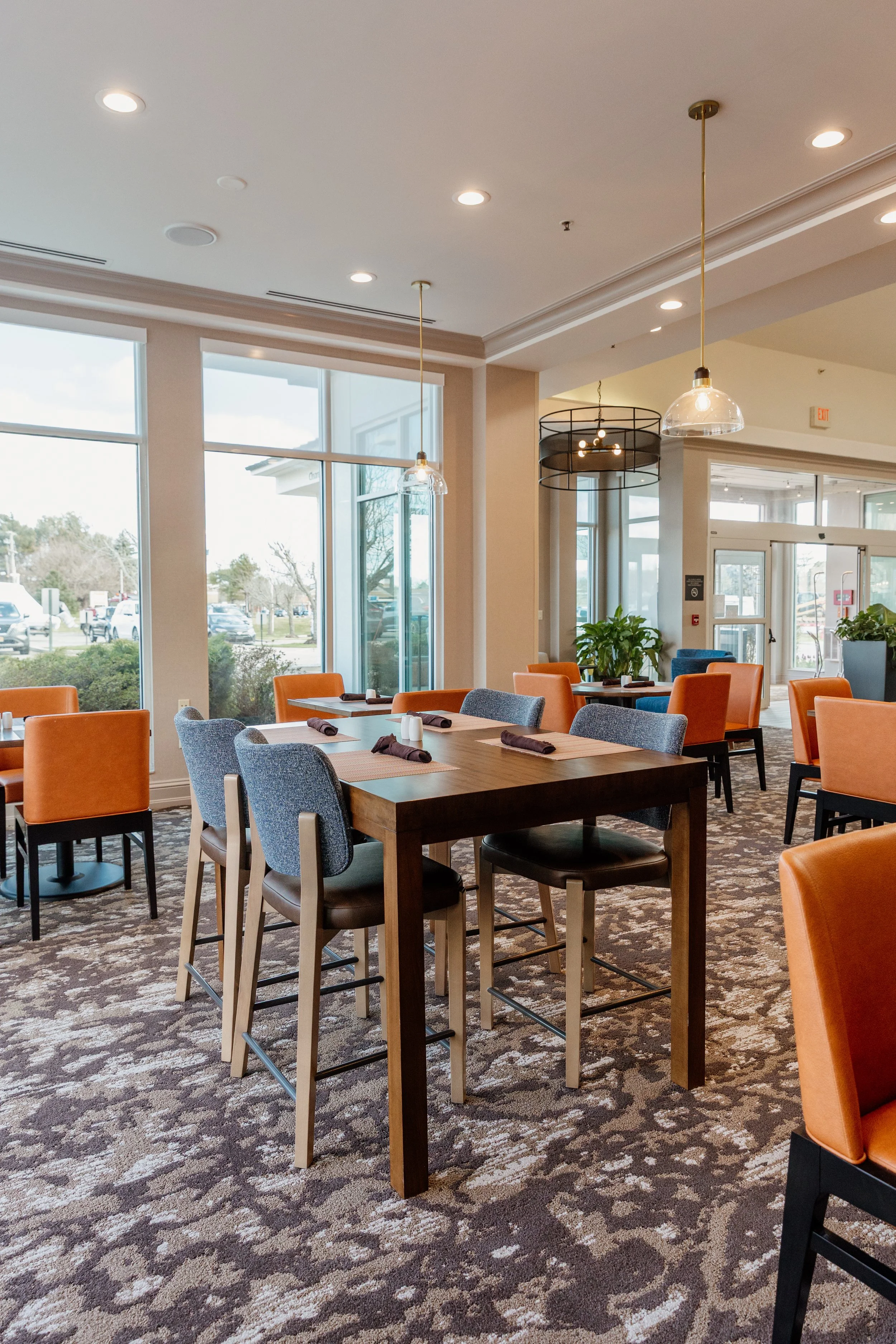 Interior of a modern restaurant or cafe with a wooden table set with napkins and salt and pepper shakers, surrounded by blue and orange chairs, large windows letting in natural light, pendant lighting, and decorative plants.