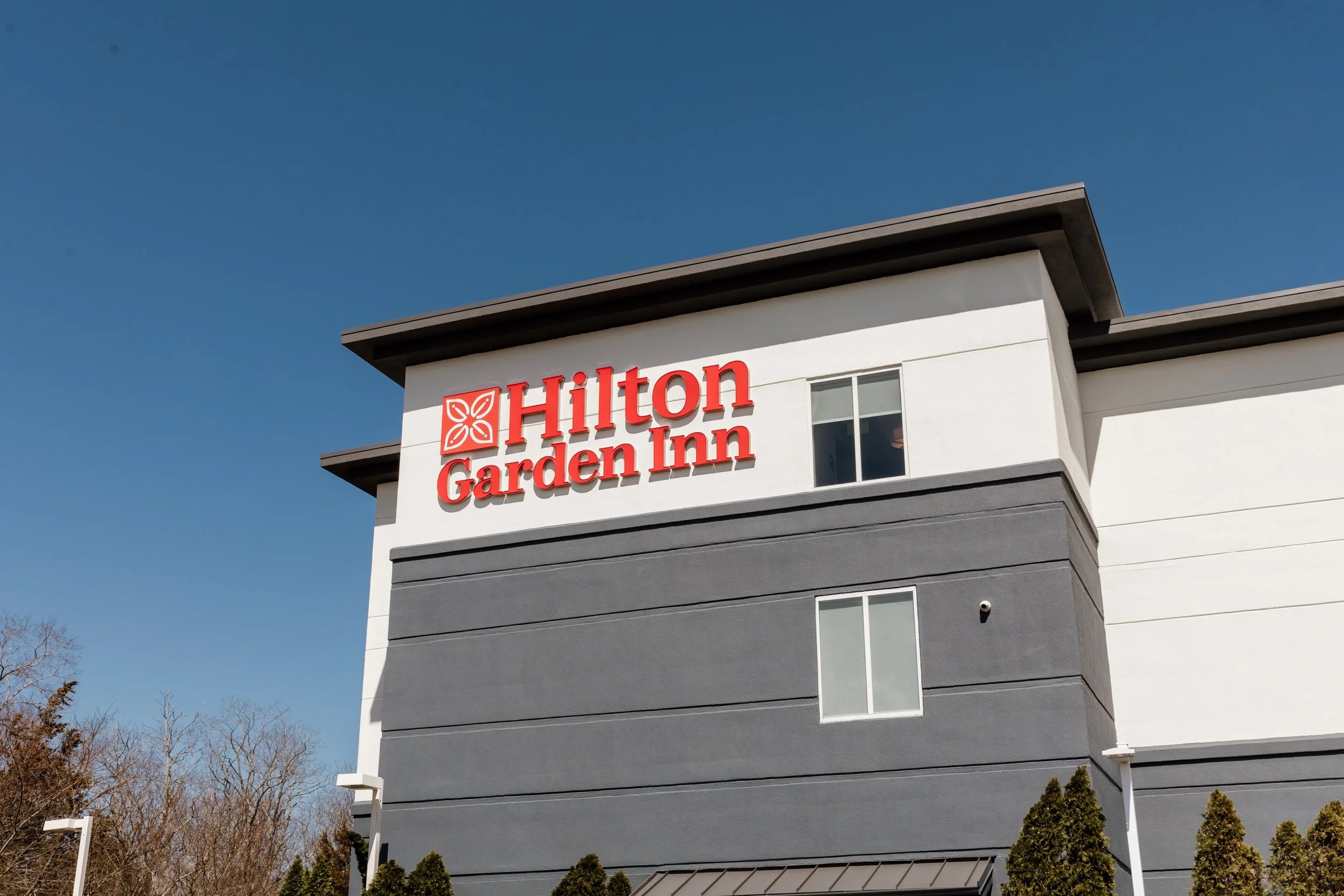 Exterior view of the Hilton Garden Inn hotel with a sign on the building, a clear blue sky, and some trees at the bottom.