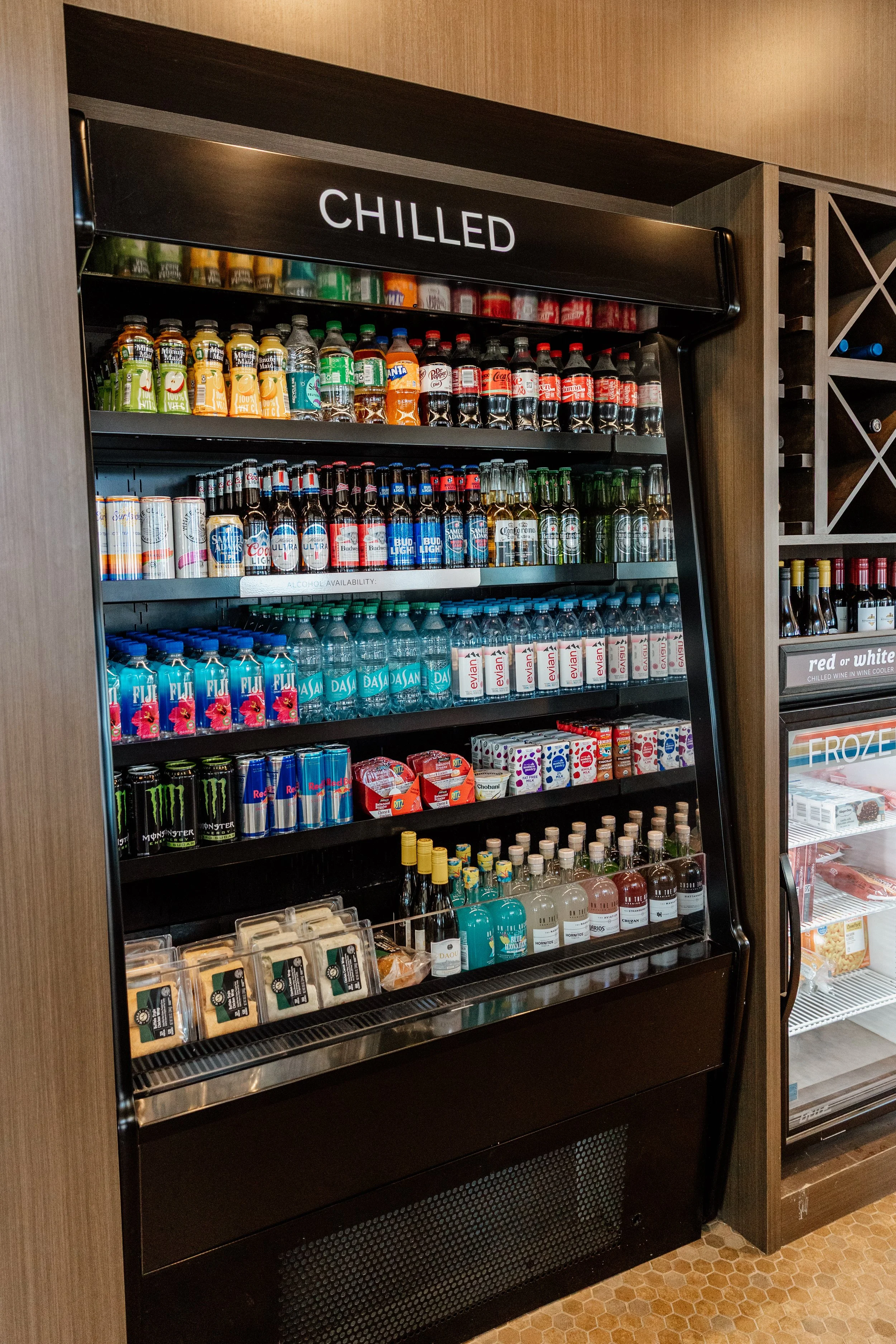 A refrigerator labeled 'Chilled' filled with various bottled and canned beverages, including juices, sodas, water, energy drinks, and alcoholic drinks, in a retail or hotel setting.