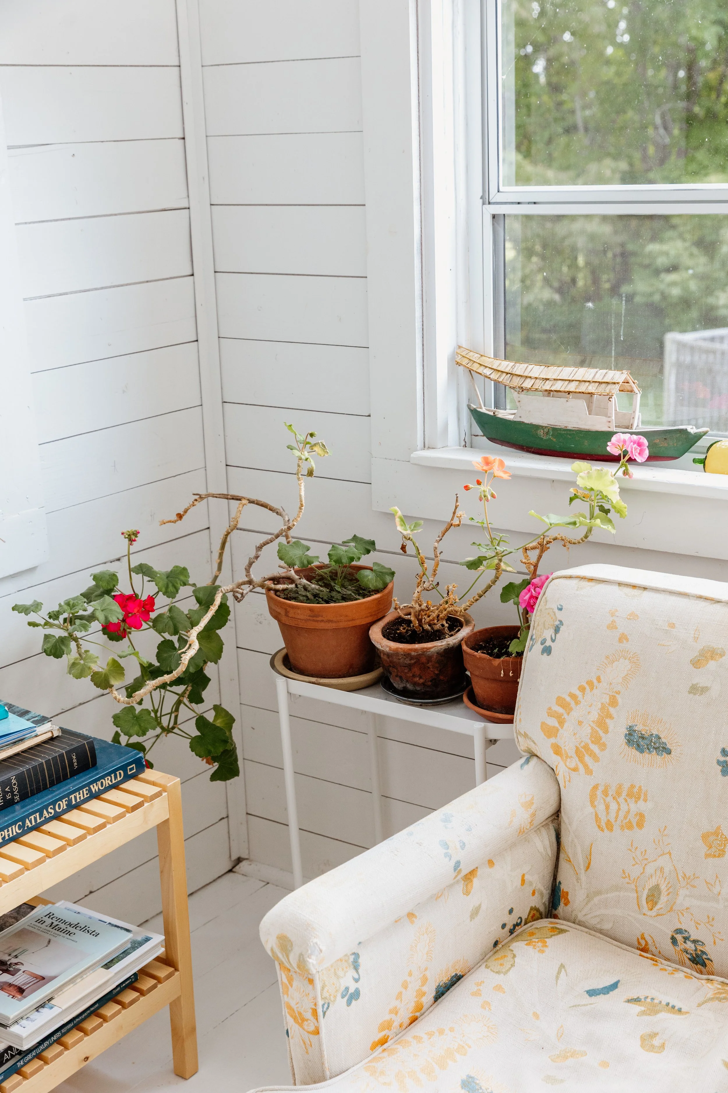 A cozy interior corner with a white wooden wall, potted plants on a small white table, a patterned armchair, a window with a boat decoration, and a bookcase with books.