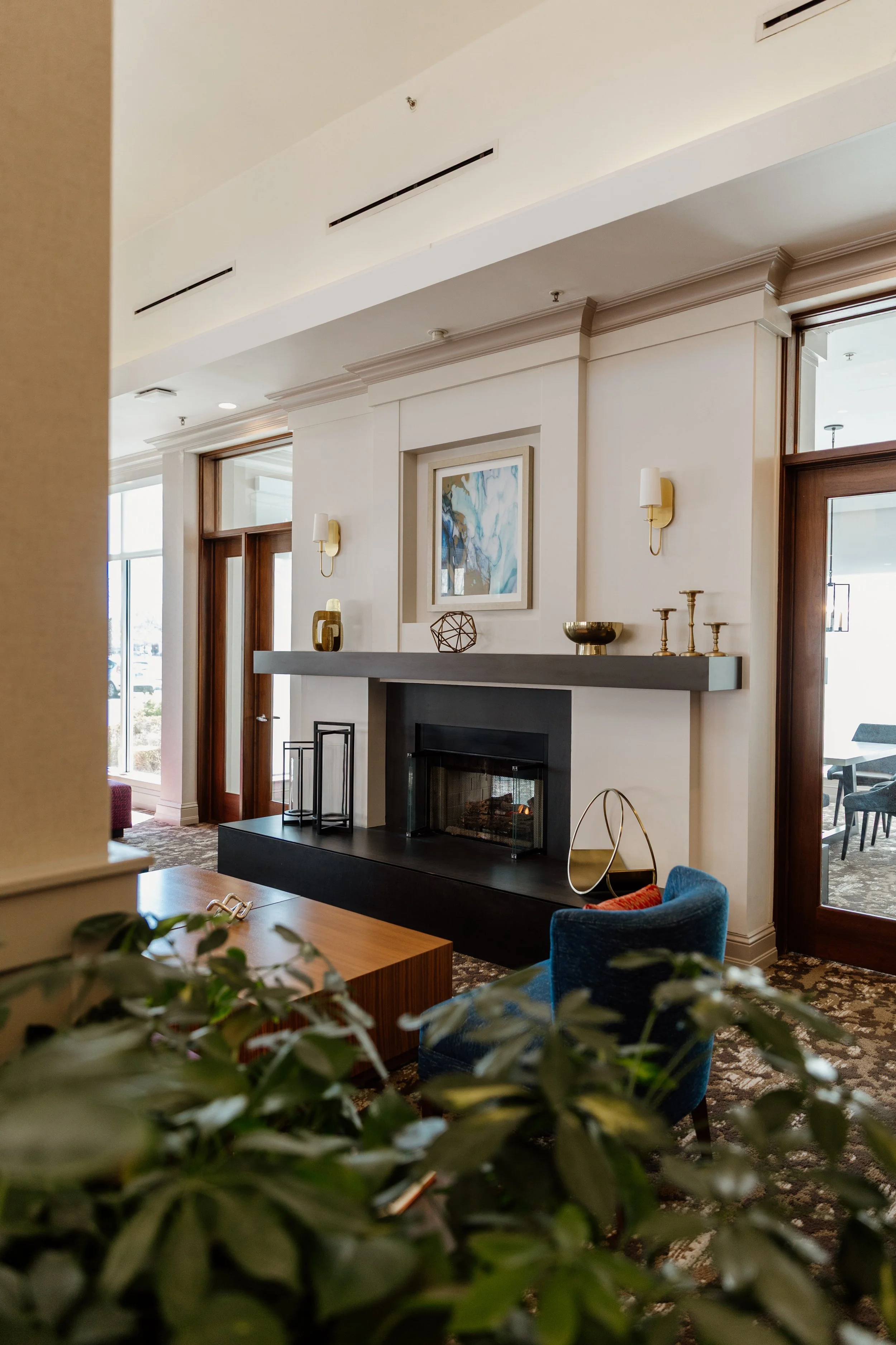 Interior of a modern living room with a white fireplace, decorative objects on the mantel, a blue upholstered chair, a wooden coffee table, and surrounding windows and doors.
