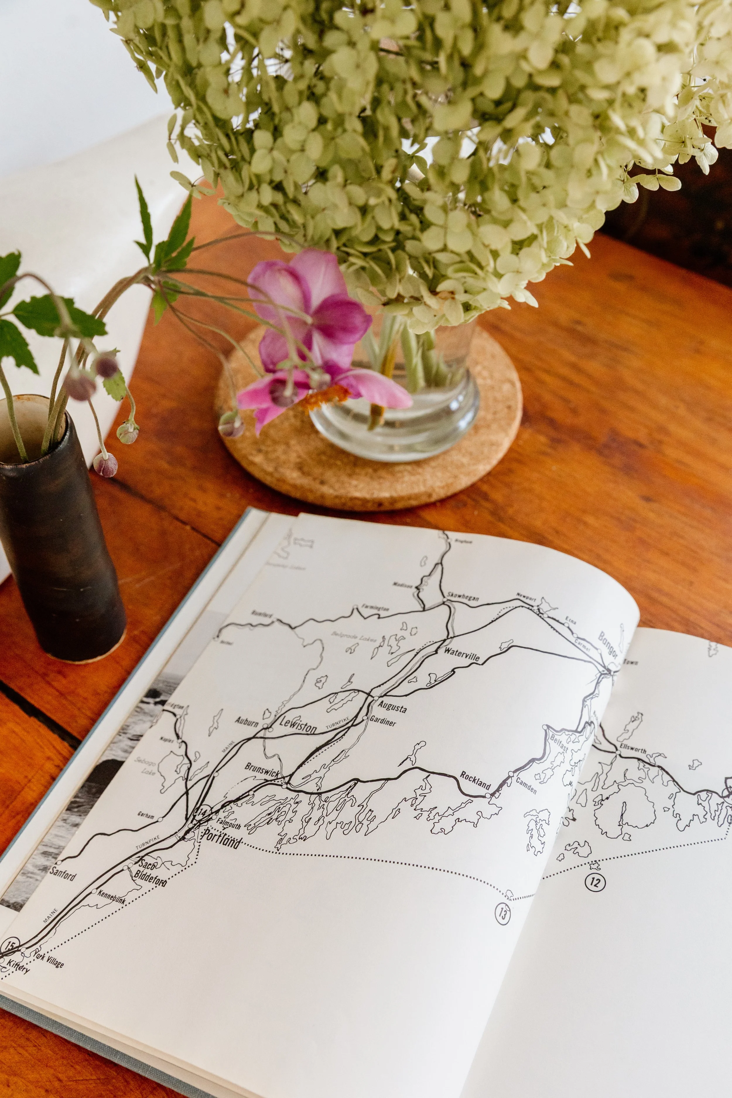 Open book showing a map laid out on a wooden table. Two vases with flowers are also on the table; one has white and green hydrangeas, and the other has pink and purple flowers.