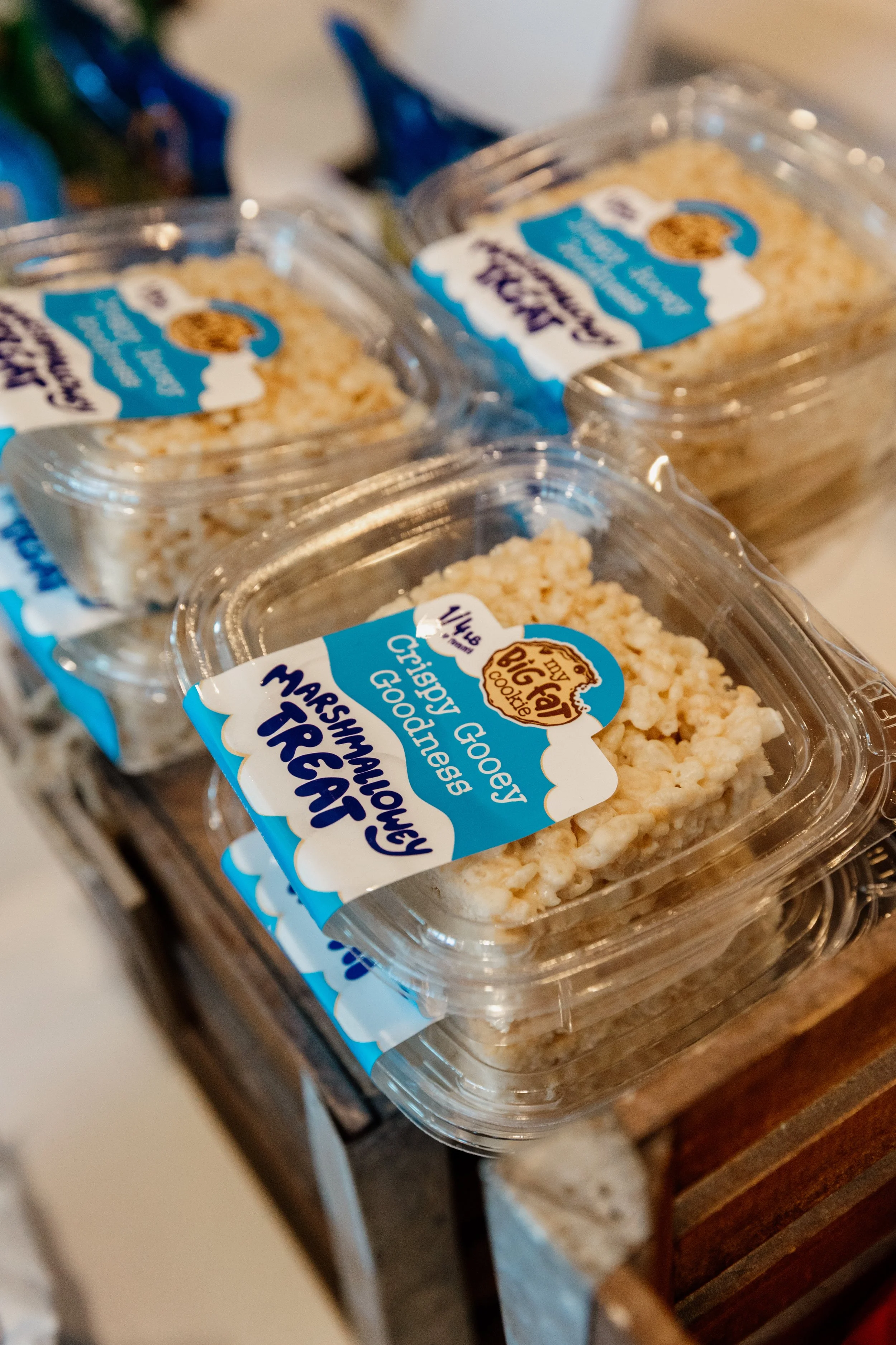 Clear plastic containers of marshmallow treats with blue and white labels reading 'Marshmallowy Treats' and 'Crunchy Goodness' stacked on a wooden surface.