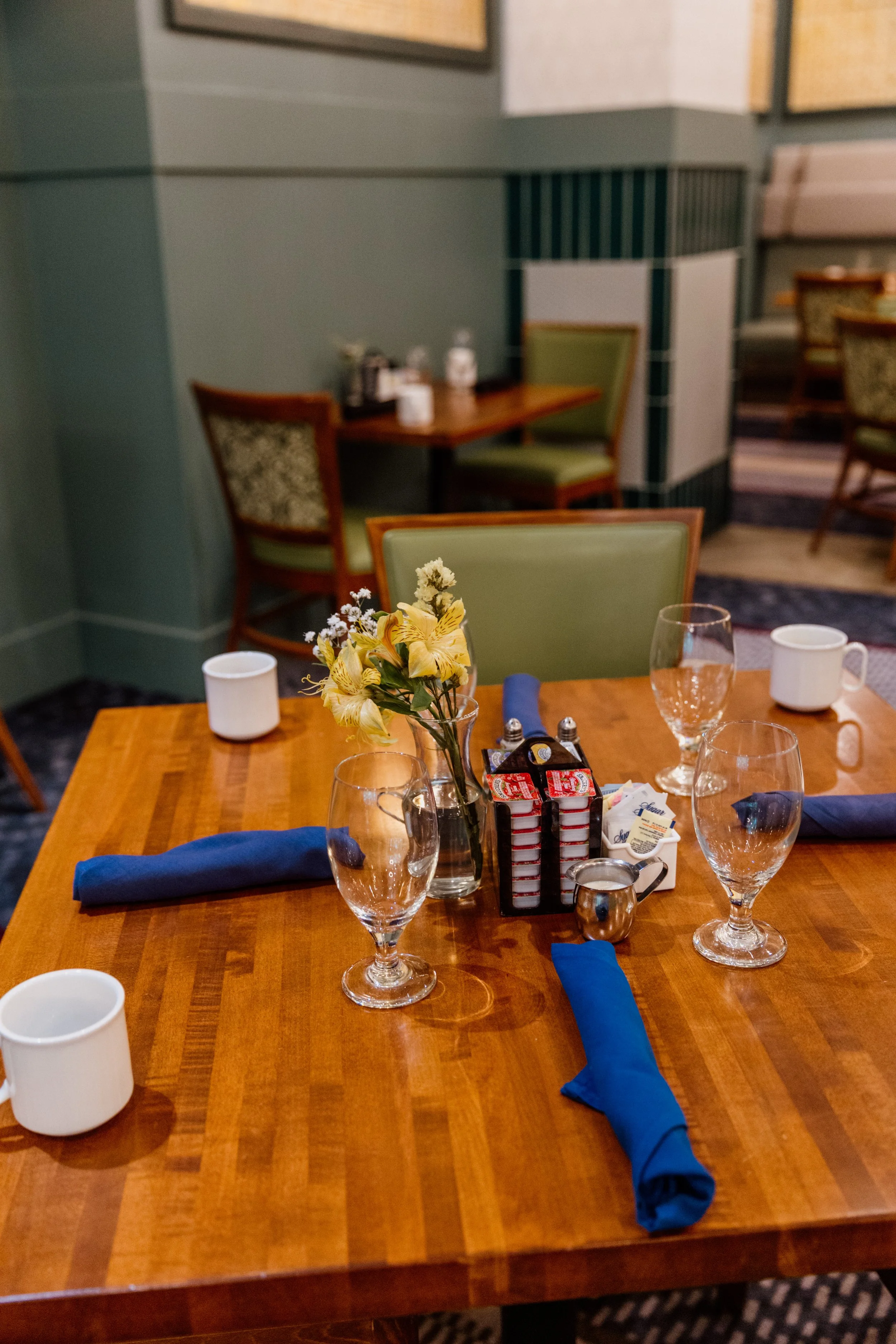 A table set for a meal with three water glasses, two white coffee mugs, blue cloth napkins, a small flower arrangement, and cream and sugar containers, in a restaurant with green chairs and wooden tables.