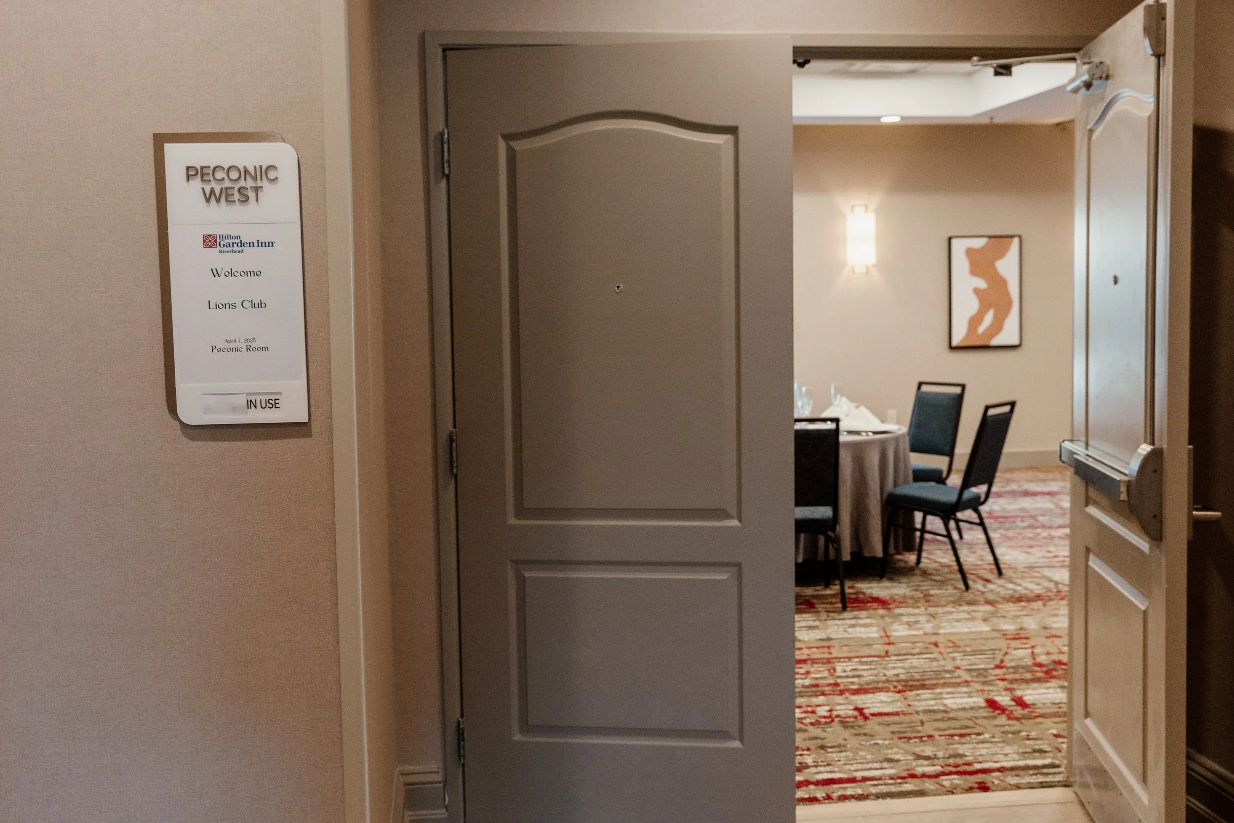 View of a hotel meeting room with a table set up, chairs, a lampshade on the wall, patterned carpet, and an abstract artwork on the wall, seen through an open door.