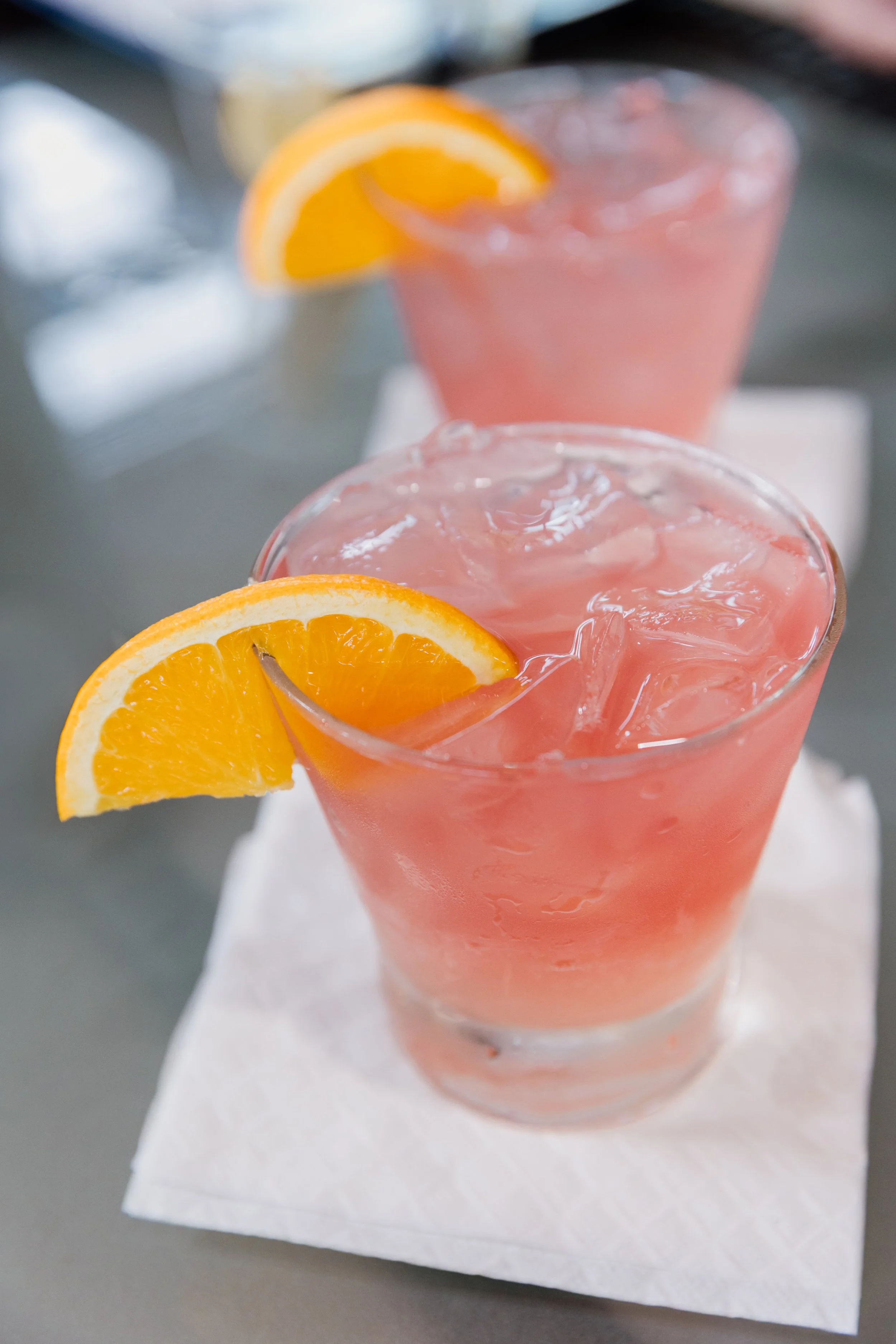 Two glasses of pink grapefruit cocktail with ice and orange slices as garnish, placed on white napkins on a gray surface.