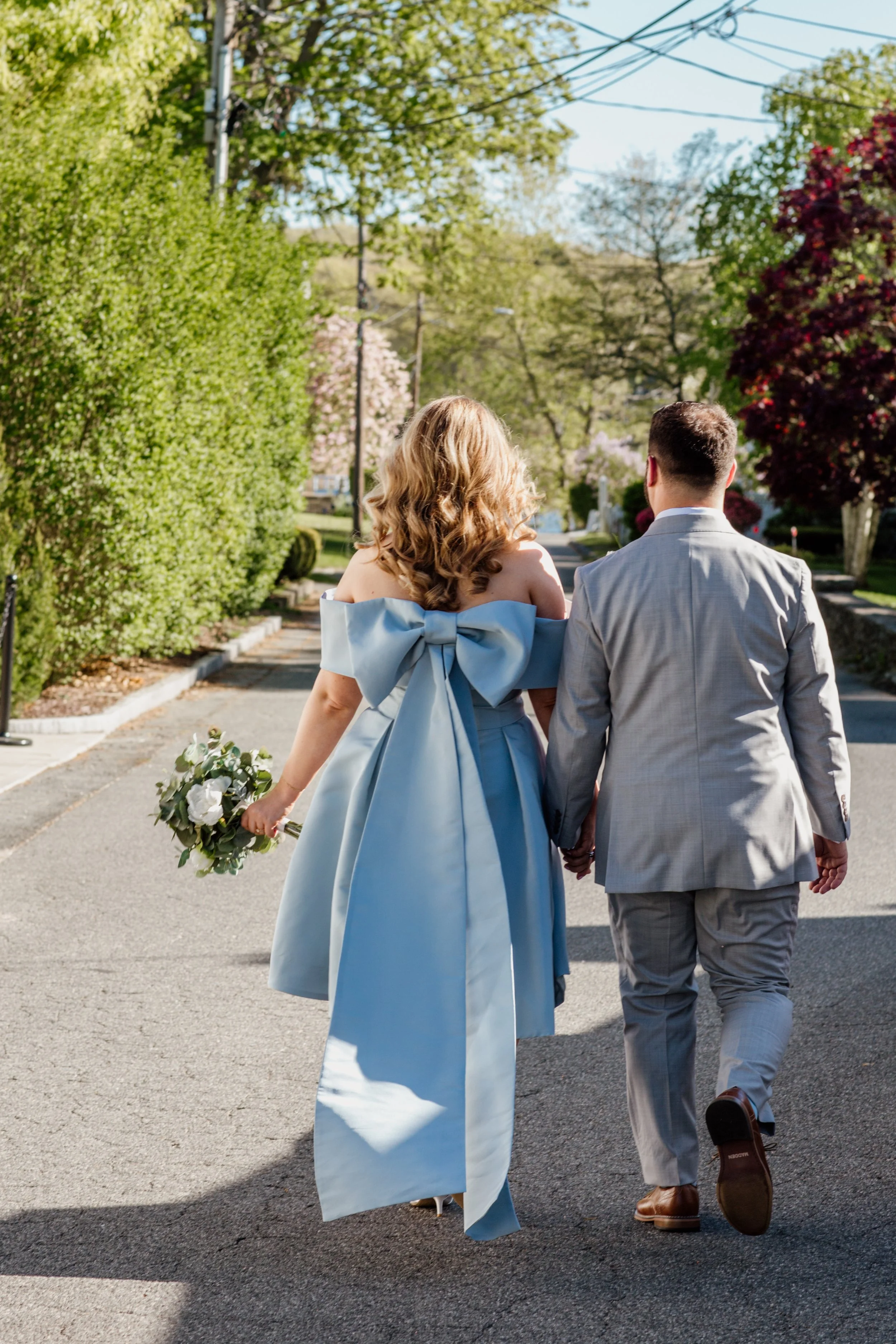 A bride in a stunning short ice blue wedding gown with a dramatic bow back walks alongside her groom in a gray suit down a sun-dappled tree-lined street in Chester, Connecticut.