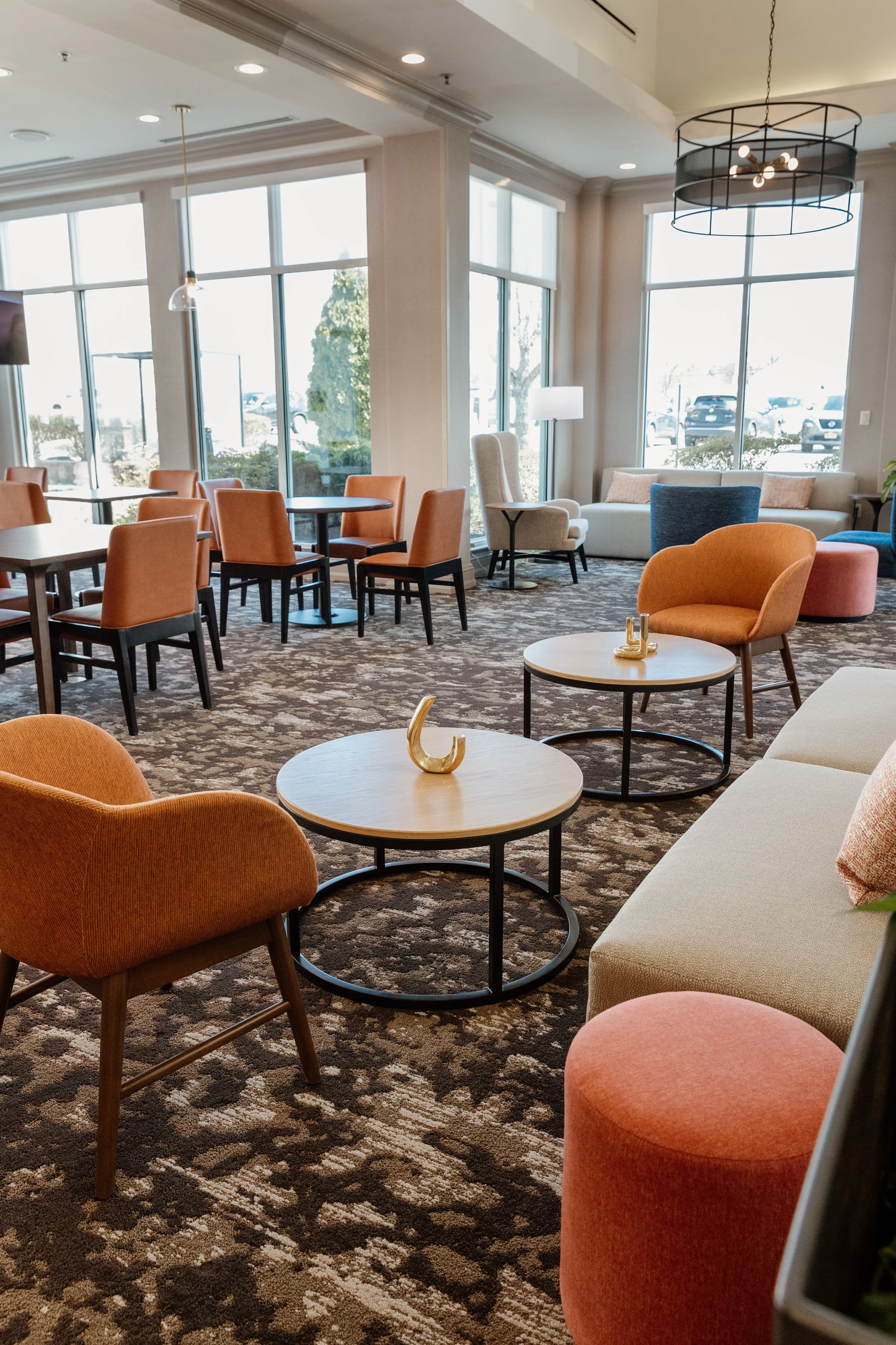 Comfortable hotel lounge with orange, blue, and beige chairs, round coffee tables, and large windows letting in natural light.