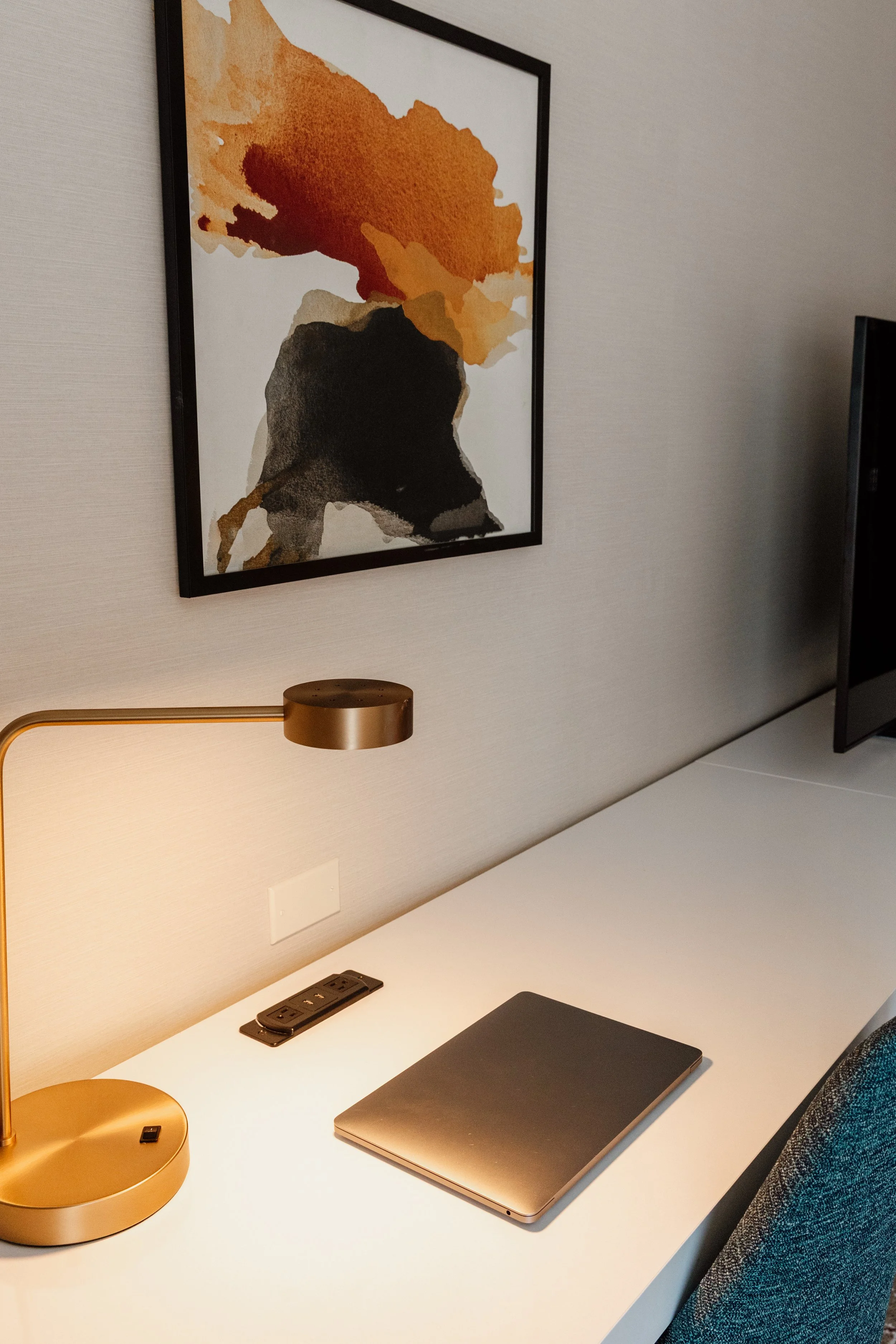 Hotel room desk with a closed MacBook, a gold-colored desk lamp, a power outlet with USB ports, and a framed abstract painting with orange, black, and gray watercolor shapes on a white wall.