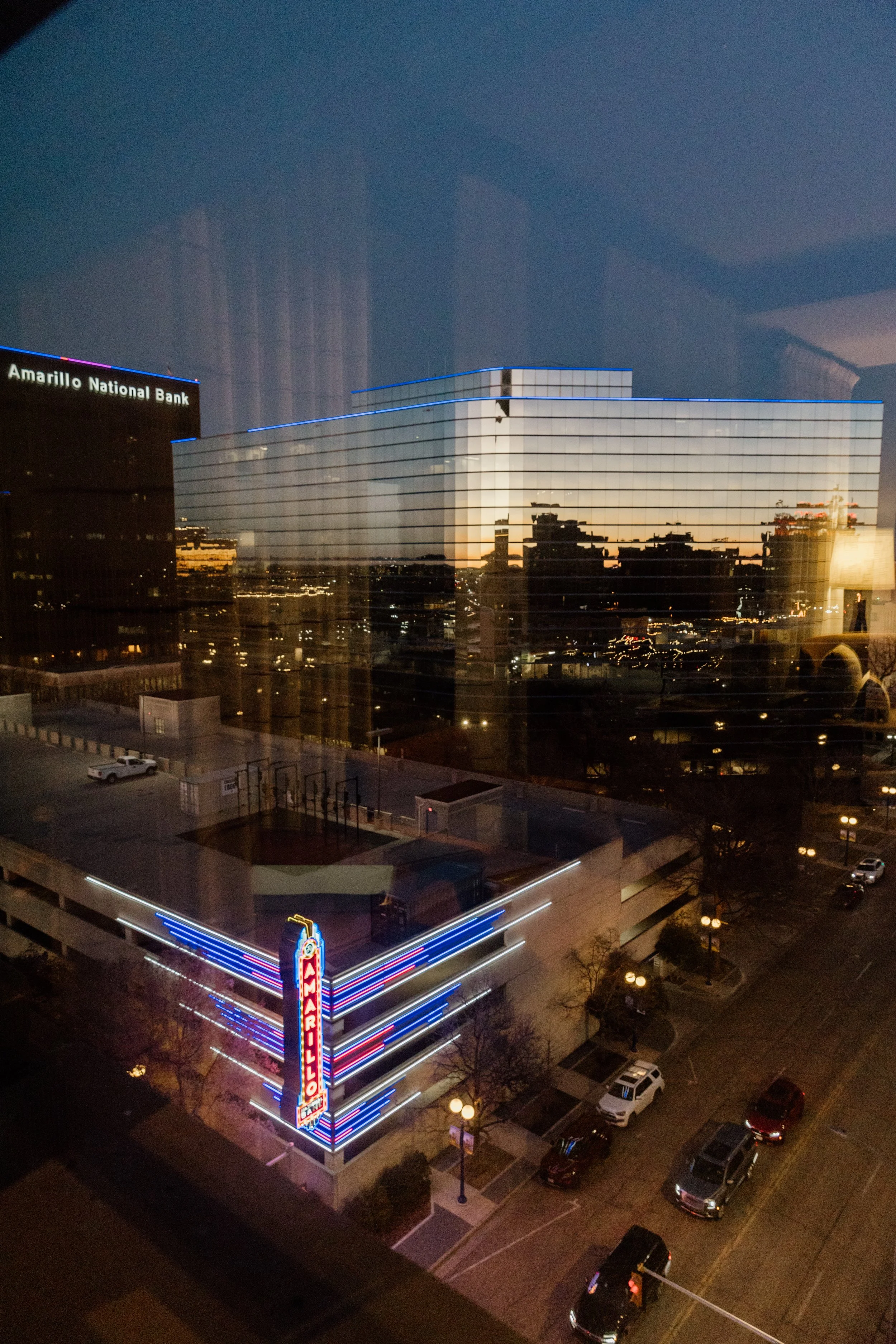 Cityscape view through a window showing reflected buildings and city lights at dusk, with the Amarillo National Bank building illuminated in neon colors in the foreground.