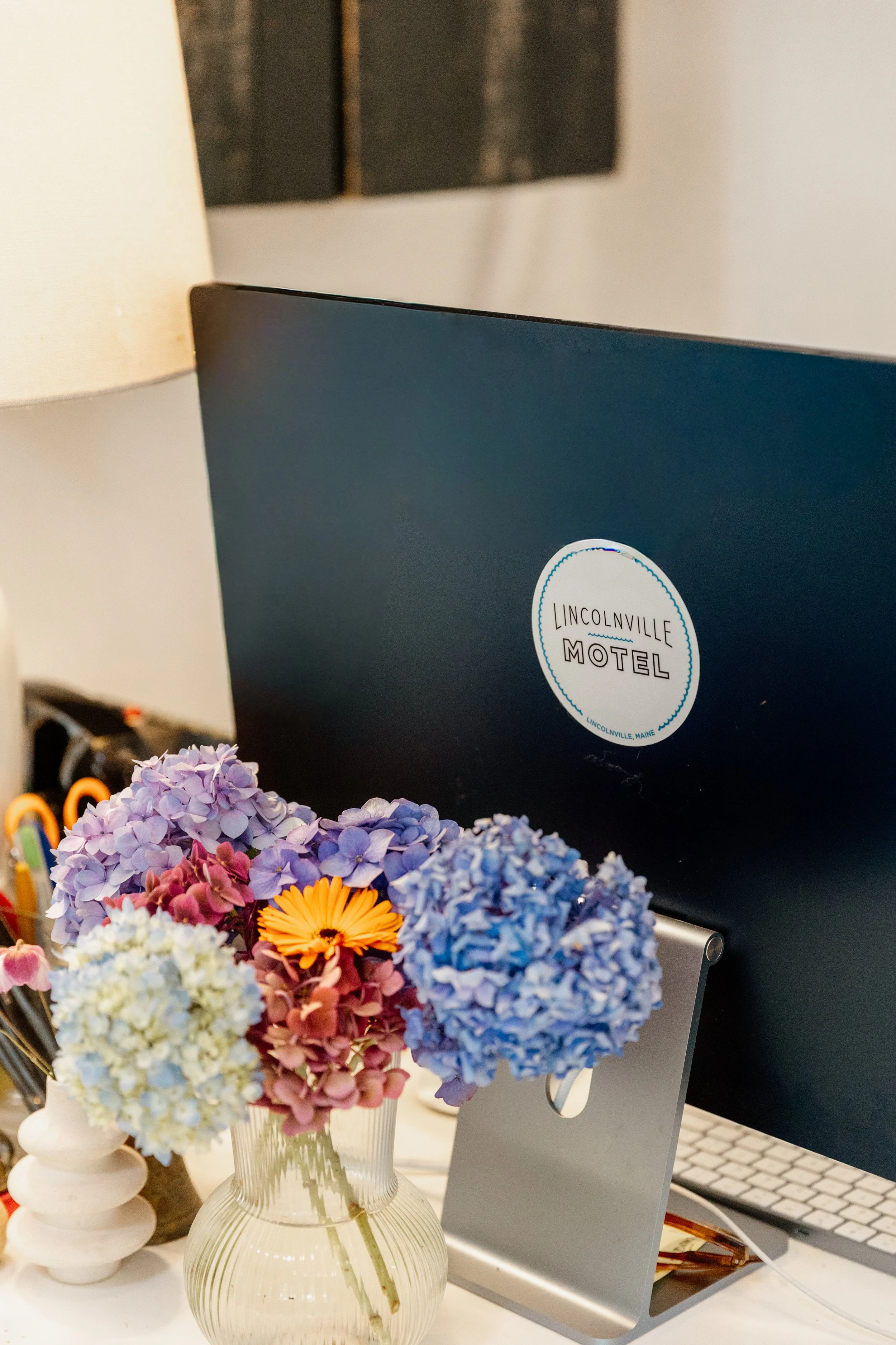 A computer monitor with a sticker that reads 'Lincolnville Motel' is placed on a desk with a glass vase of colorful hydrangeas flowers in front of it.