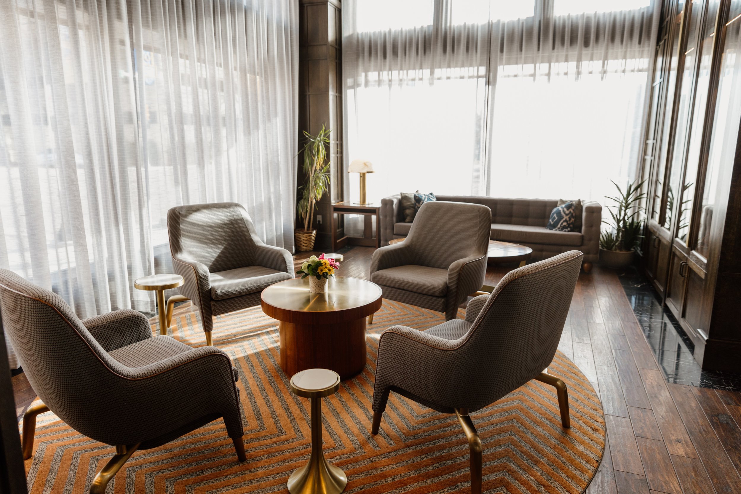 A warm and inviting hotel lobby seating area with armchairs, a round wooden coffee table, fresh florals, and sheer curtained windows, captured during a content business shoot at the Graduate by Hilton in Fayetteville, Arkansas.