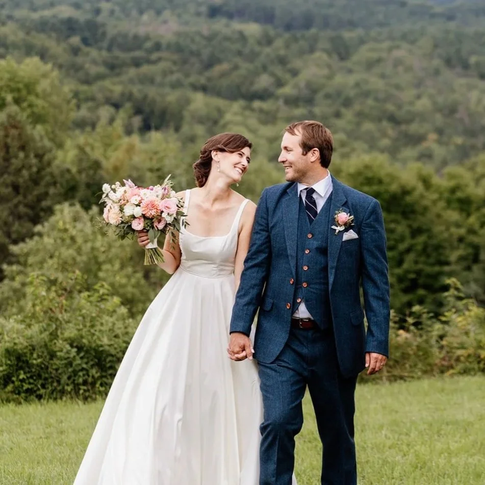 A bride in a white wedding dress holding a bouquet of pink and white flowers walks hand-in-hand with a groom in a blue suit with a pink boutonniere in a lush green outdoor setting.