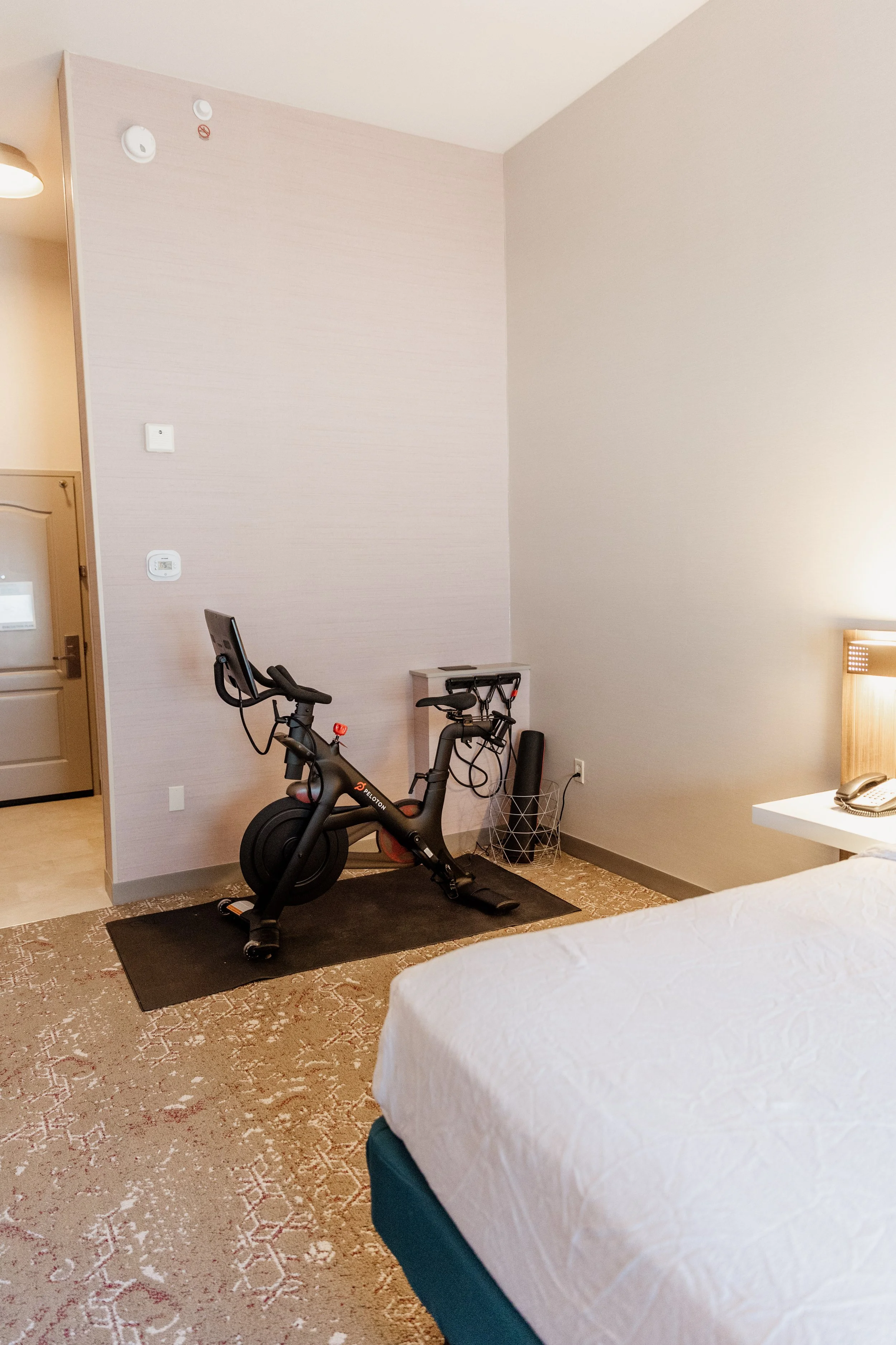 A hotel room with a stationary exercise bike on a black mat, a portion of a bed with white bedding, a nightstand with a phone, and a wall with a smoke detector and thermostat.