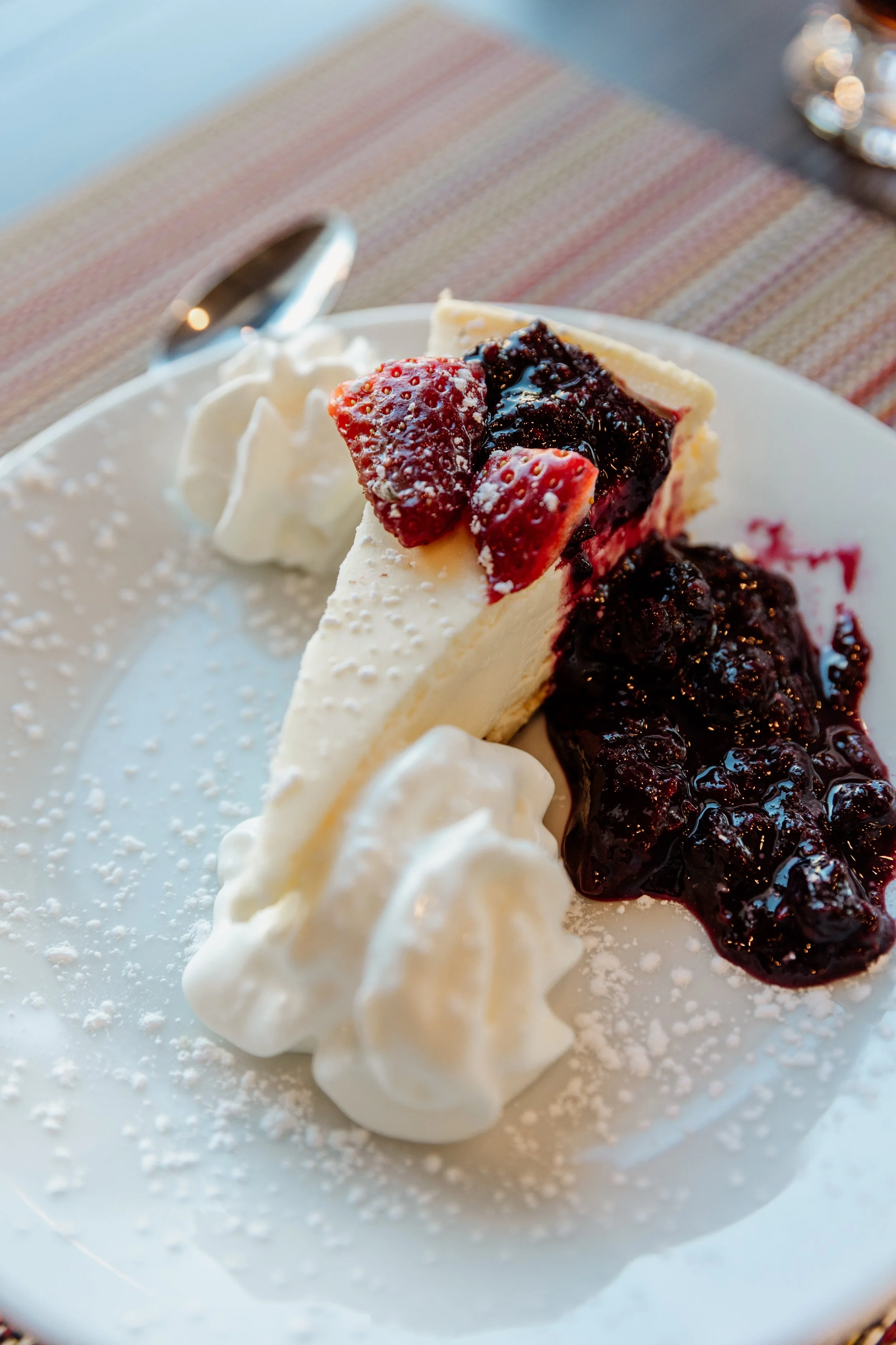 A slice of cheesecake topped with strawberries and berry compote, served with whipped cream on a white plate.
