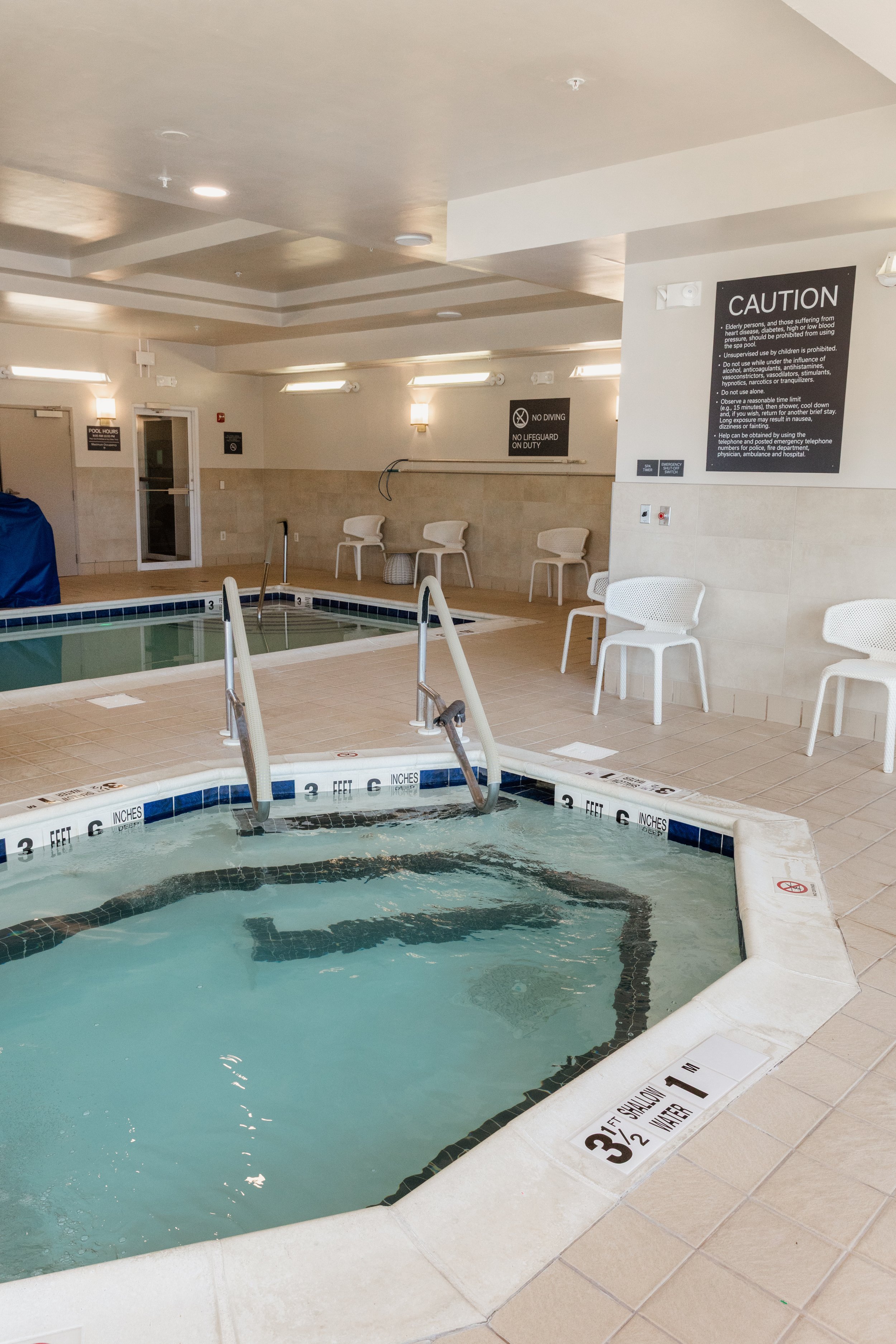 Indoor pool area with two hot tubs, white chairs lining the walls, and safety signs on the wall including 'No Diving' and 'Caution' notices.