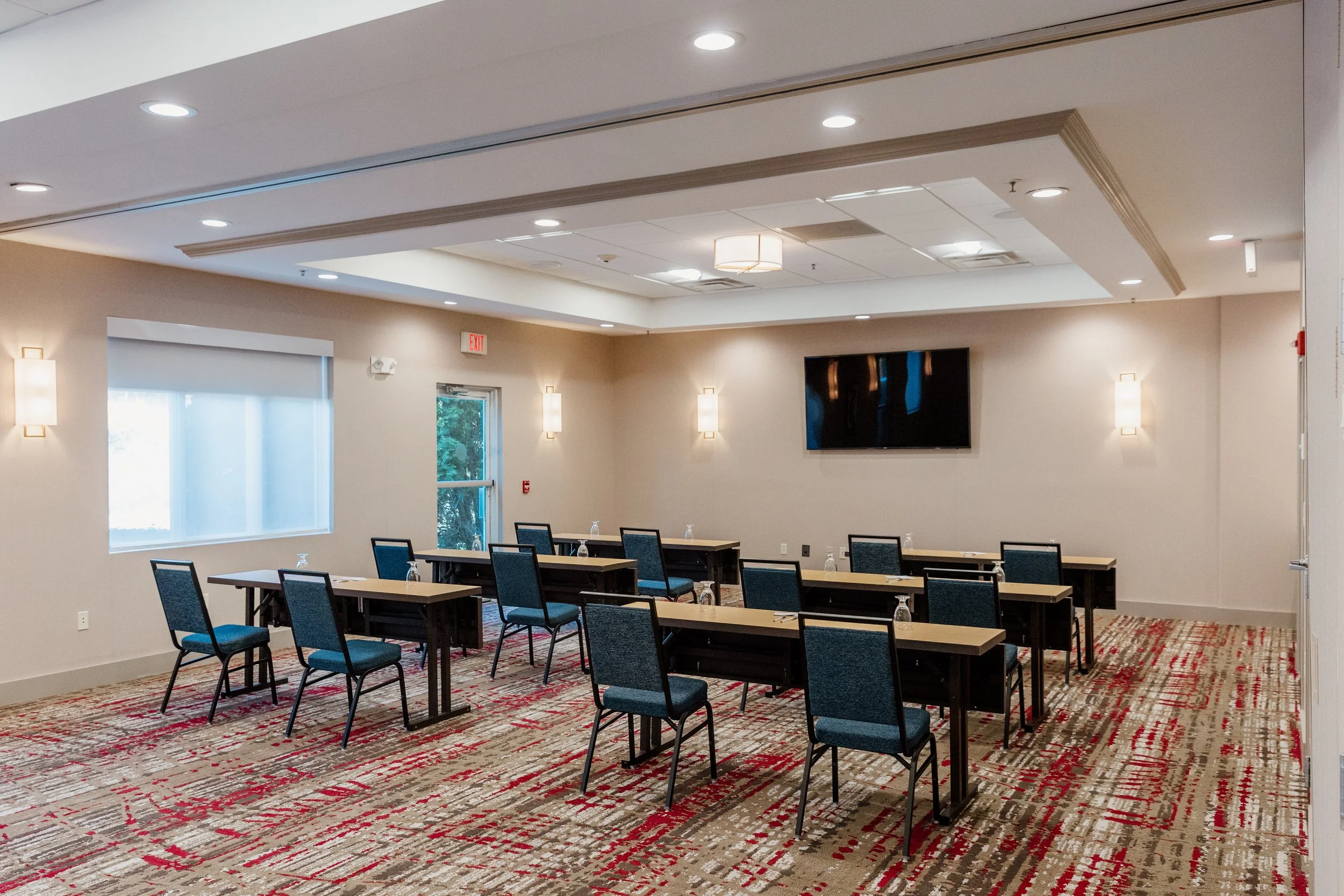 A conference room arranged with nine tables and chairs, a wall-mounted TV, a window, and a door with blinds, featuring beige walls, modern lighting, and patterned carpet.