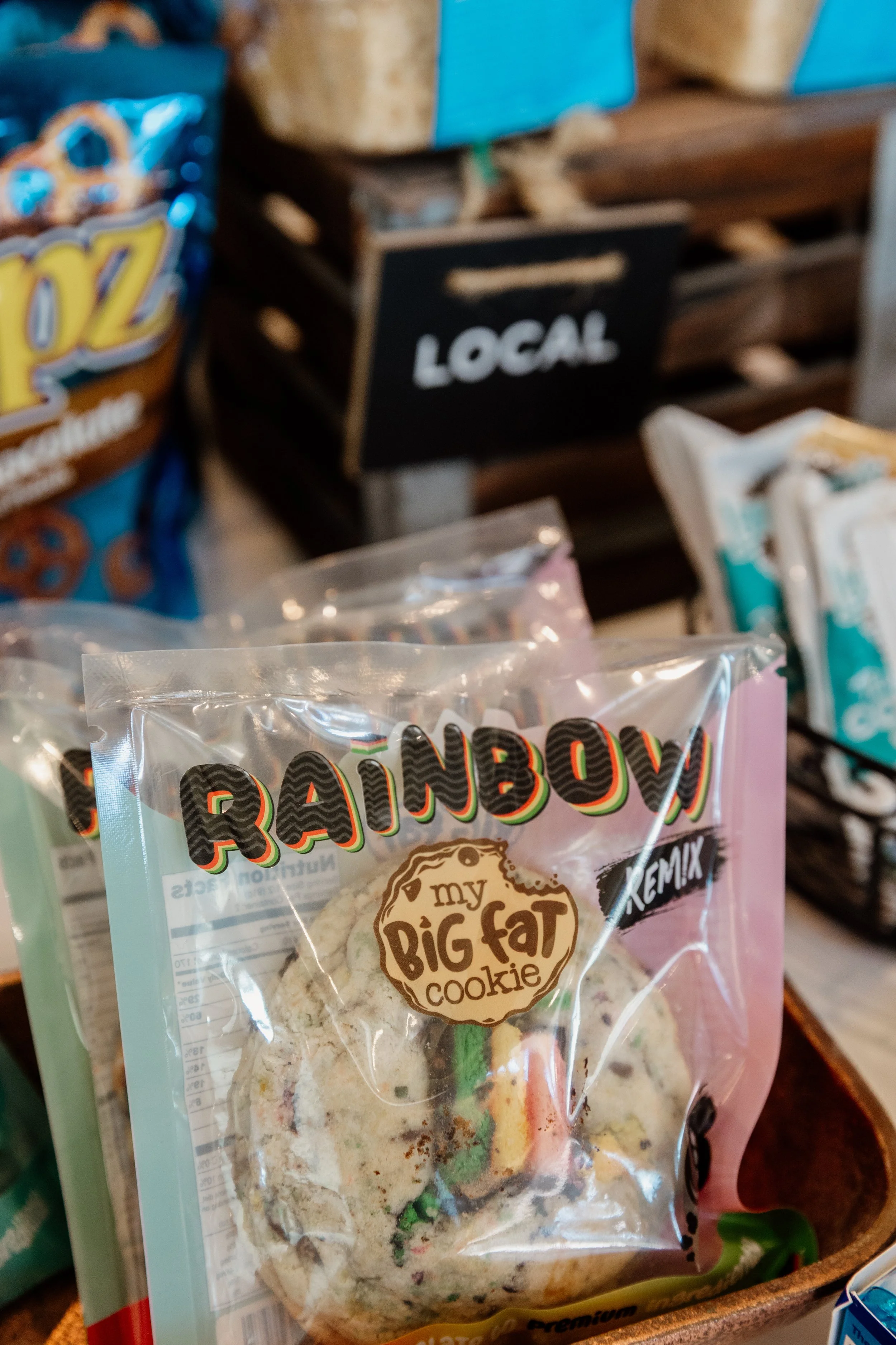 Close-up of a rainbow cookies bag labeled "my BIG fat cookie" on a grocery store shelf with other snack packages and a black "LOCAL" sign in the background.