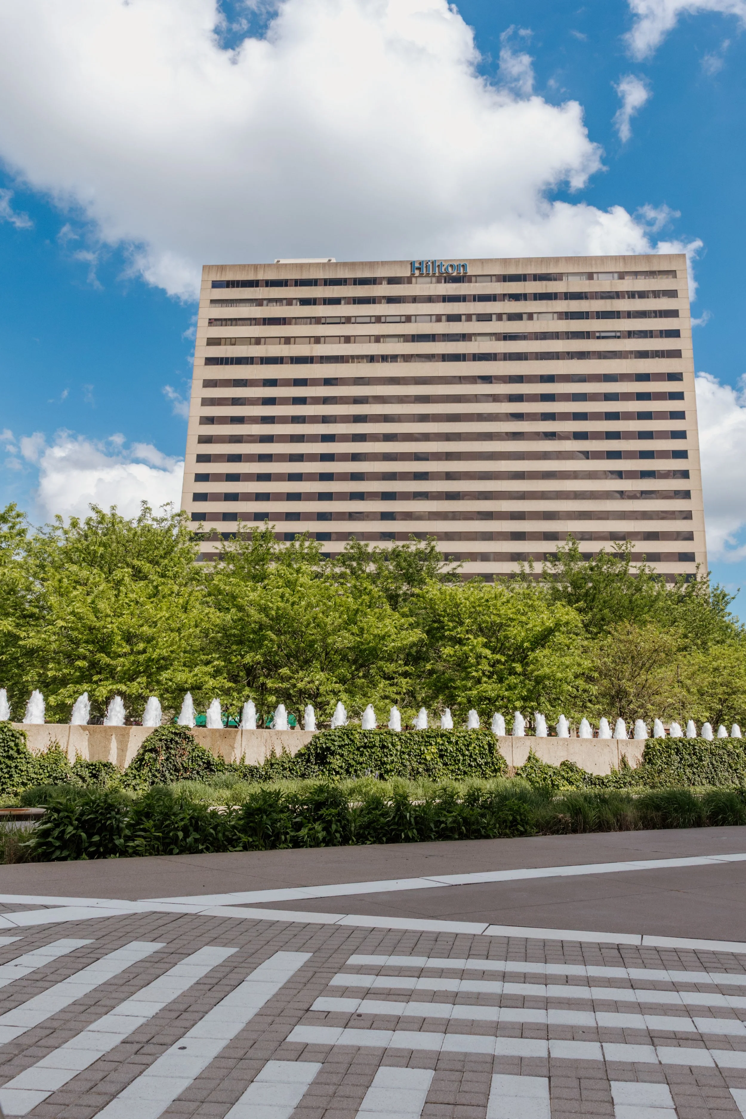 A tall beige Hilton hotel building with numerous windows under a partly cloudy sky, with green trees, fountains, and a decorative tiled walkway in the foreground.