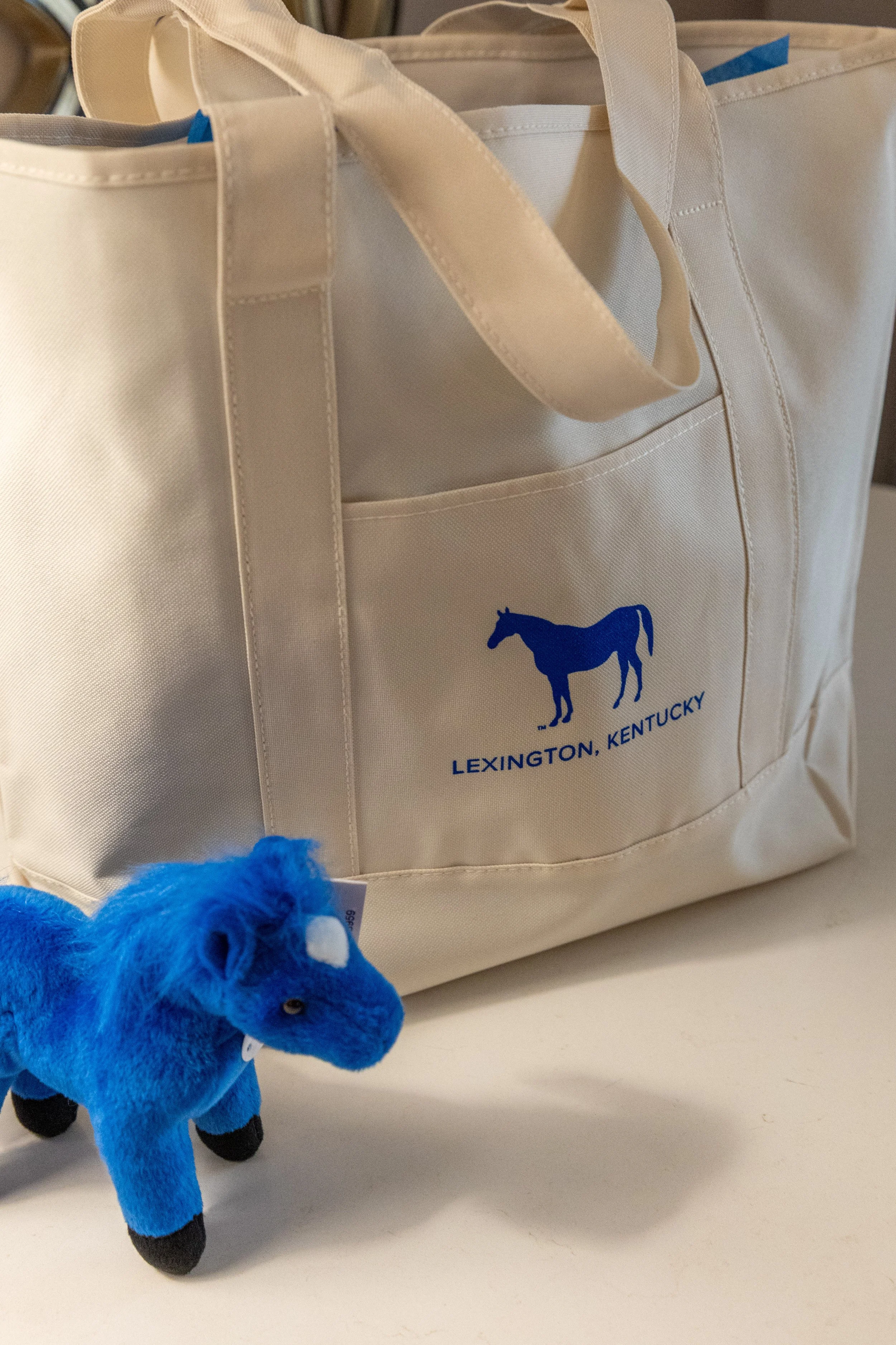 White tote bag with a blue horse logo and the words "Lexington, Kentucky" printed on it, placed on a surface. In front of the bag, there is a small blue stuffed horse toy with black legs.
