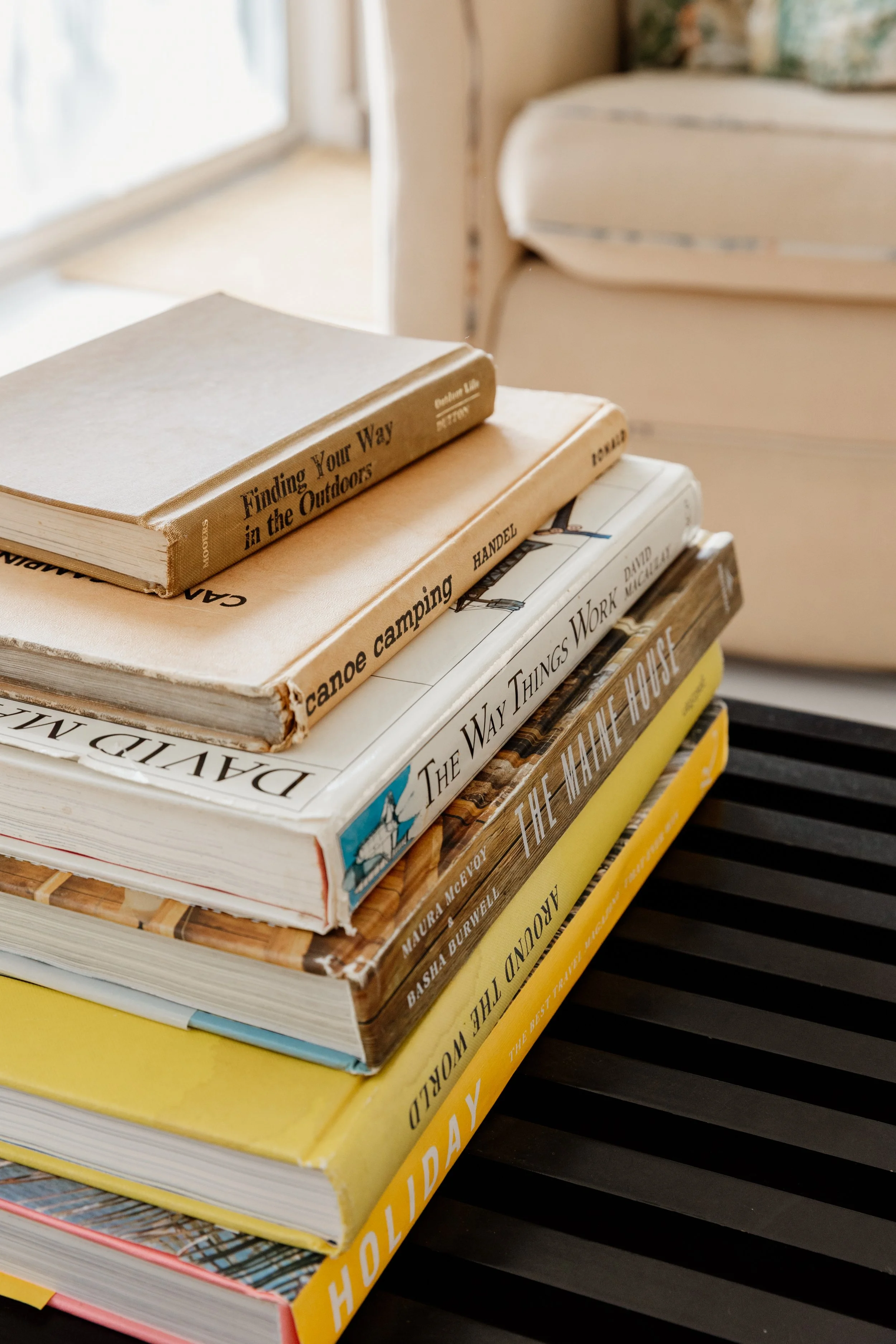 Stack of hardcover and paperback books on a dark surface near a window, with a bed in the background