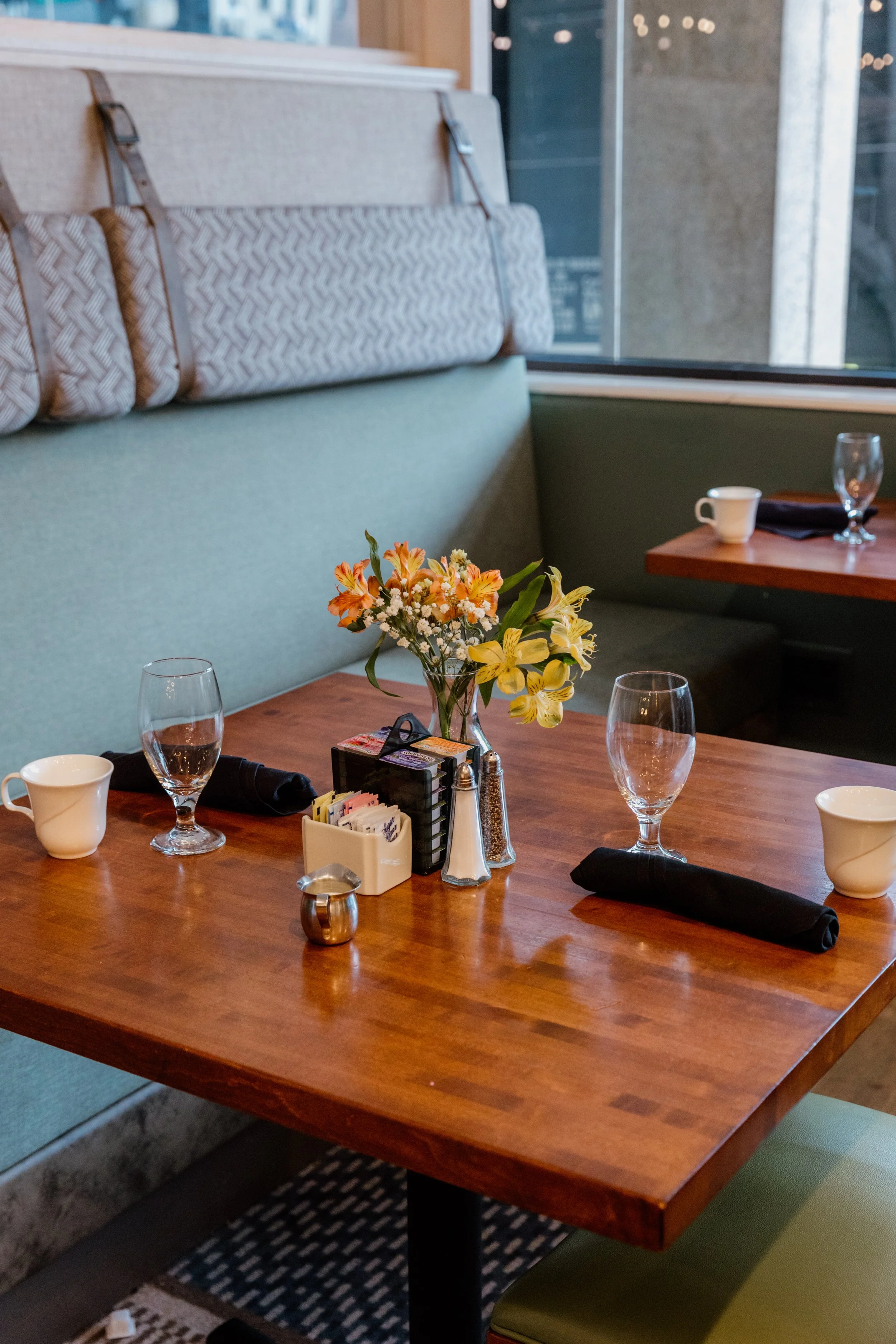 A restaurant table set for two, with a small flower arrangement, salt and pepper shakers, coffee mugs, empty wine glasses, and black napkins. The table is near a window with a view of a building exterior.