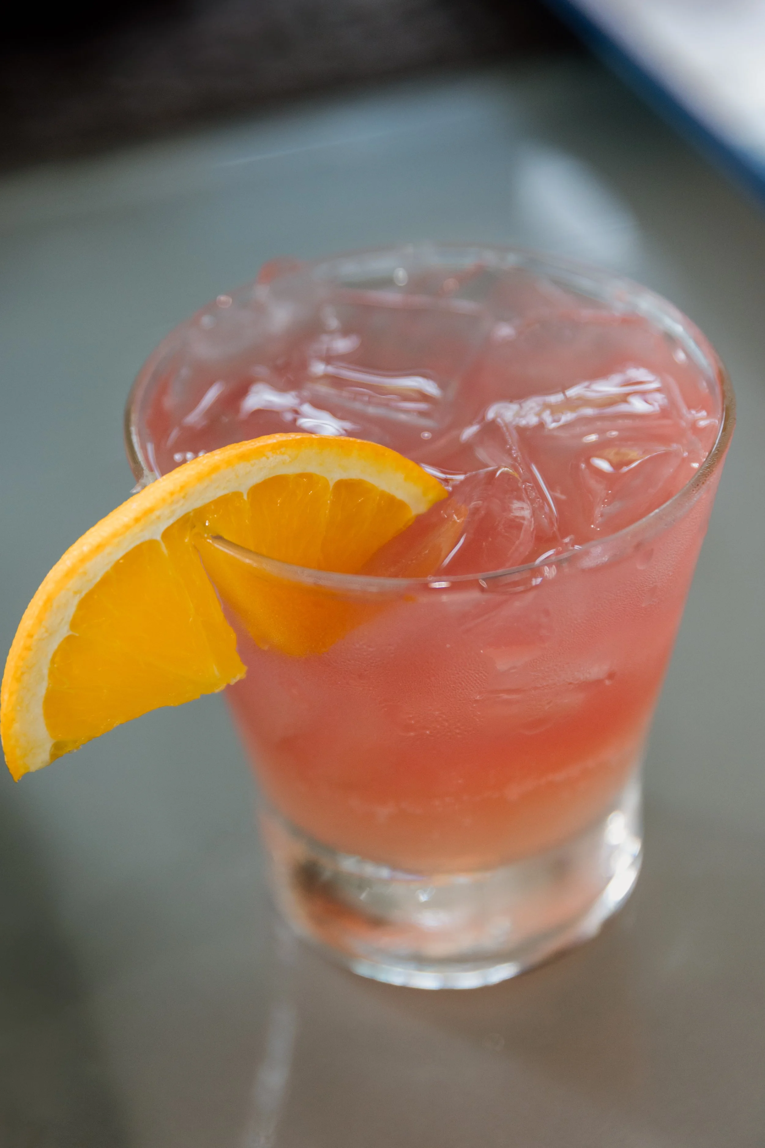 A glass of pink-colored beverage with ice cubes and a slice of orange on the rim, on a gray surface.
