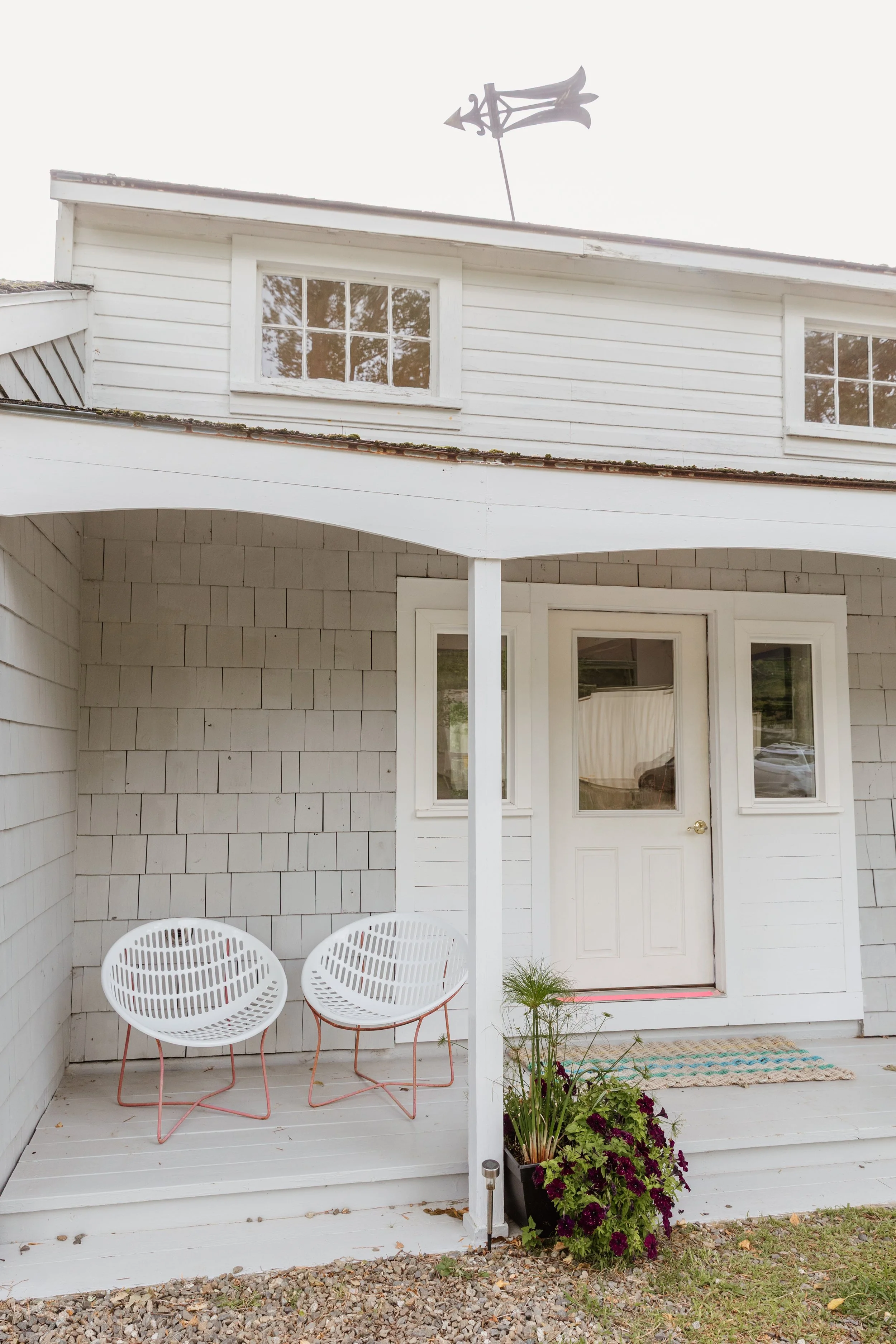 Front porch of a white house with two modern white chairs, a door with glass panel, plant in a pot, and a decorative weather vane on the roof.