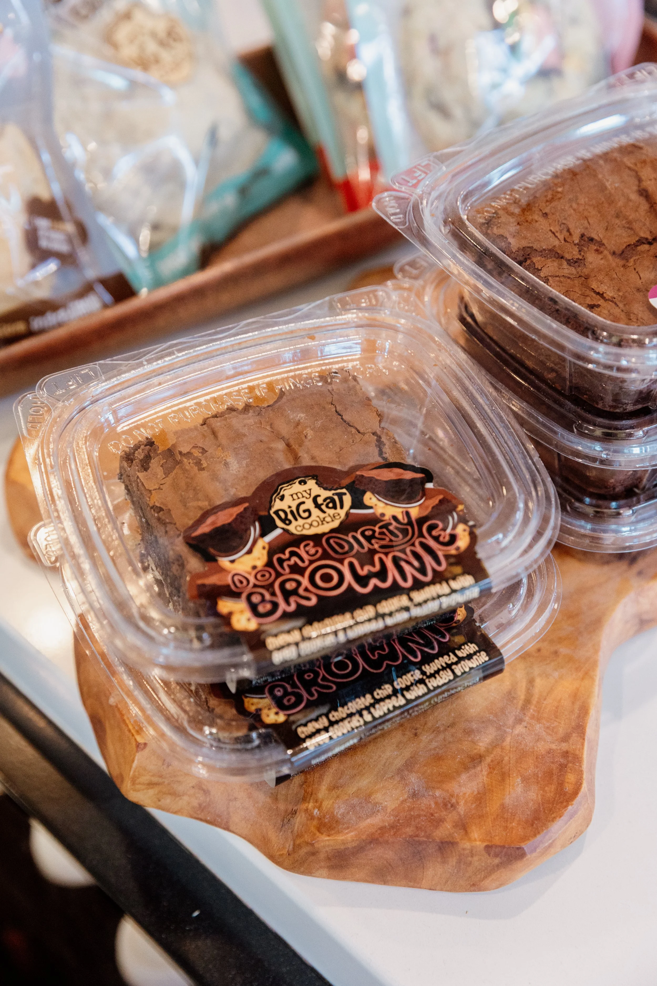 Clear plastic containers of chocolate cookies labeled "Big Fat Chocolate Brownie" on a wooden tray, with other packaged baked goods in the background.