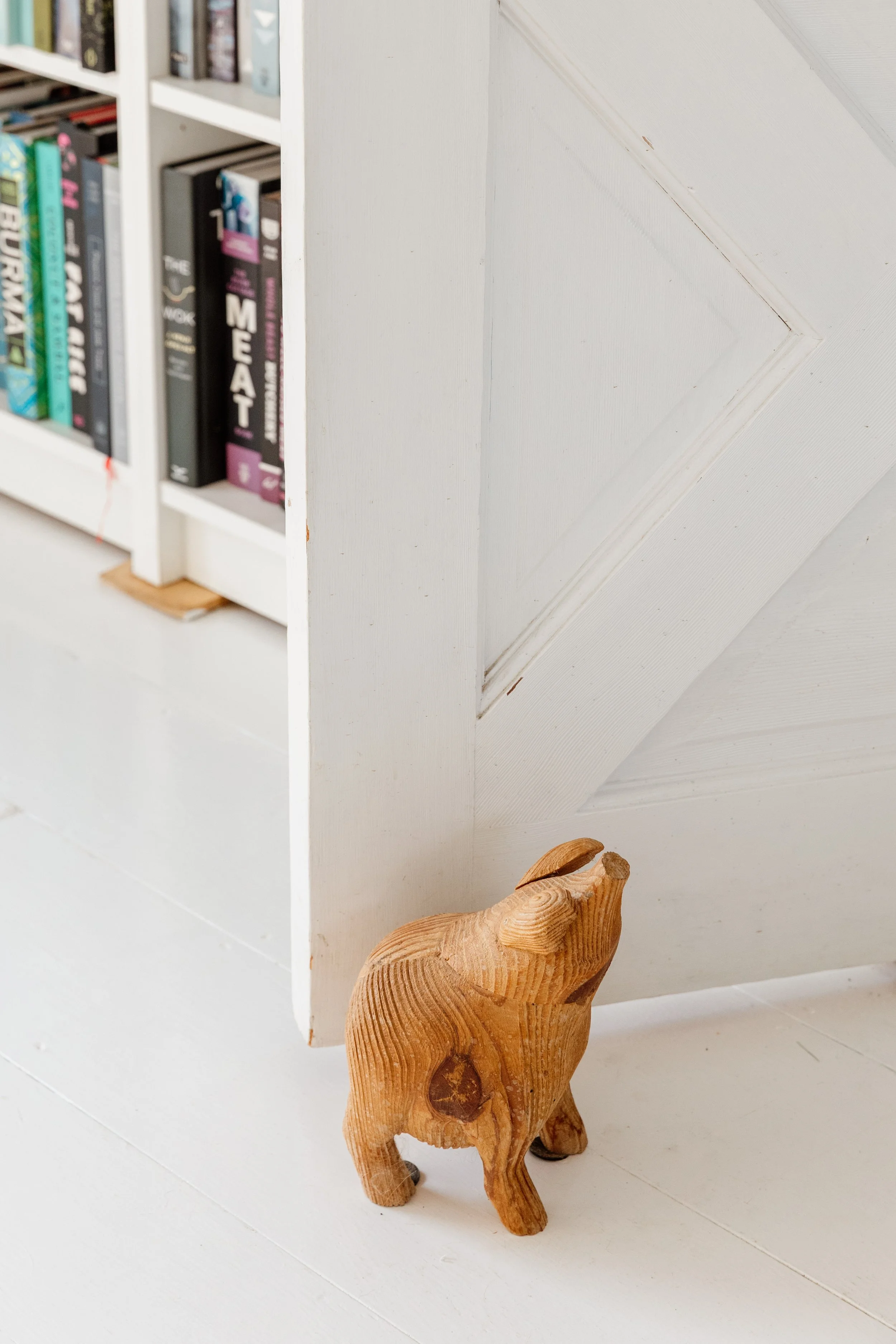 Wooden elephant figurine on a white floor next to a white cabinet or wall, with part of a bookshelf visible.