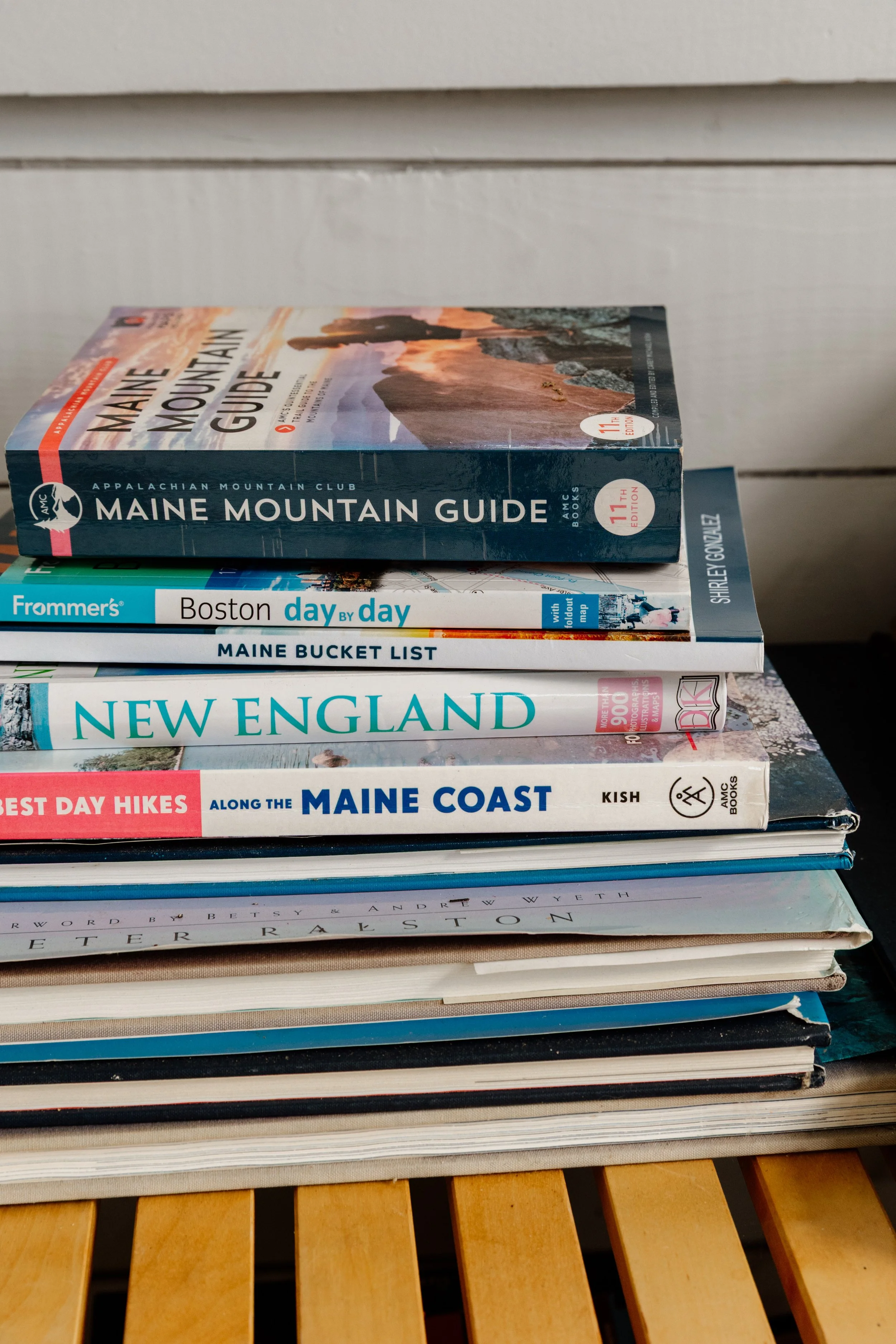 Stack of travel guides and books on a wooden surface, including Maine Mountain Guide, Boston day by day, Maine Bucket List, New England, and along the Maine coast.