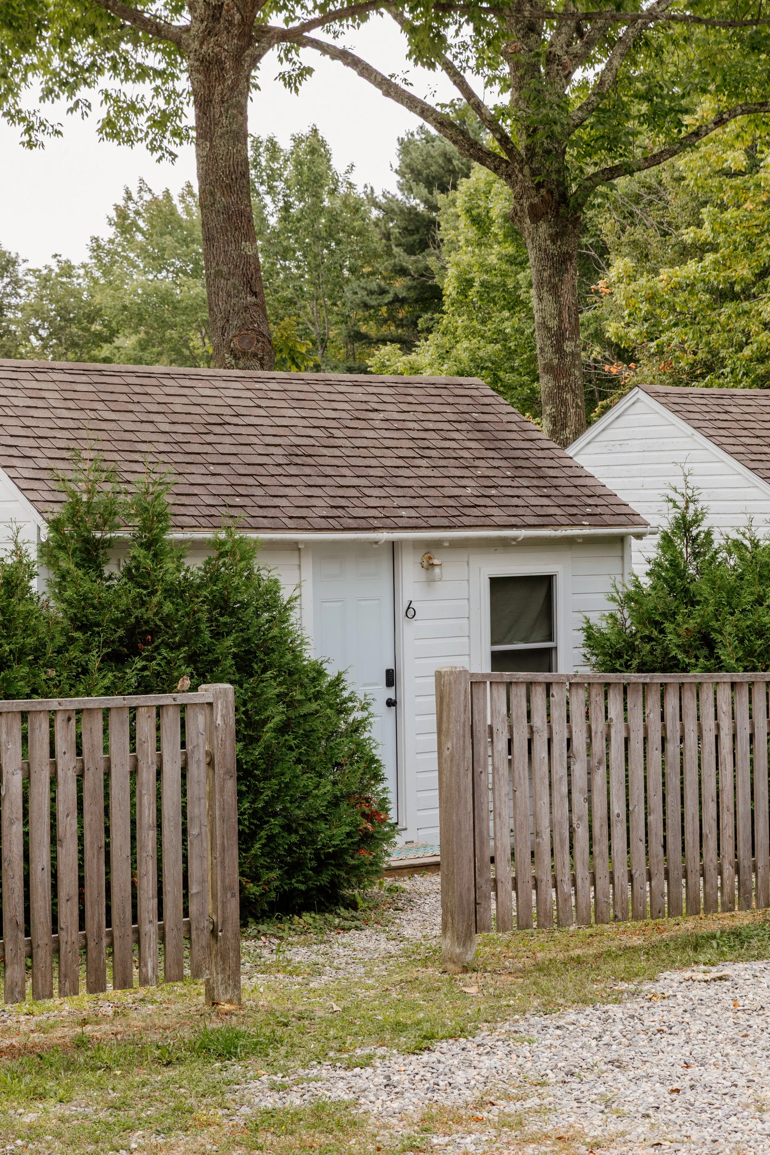 A small white house with a brown shingled roof behind a wooden fence, surrounded by green bushes and trees.
