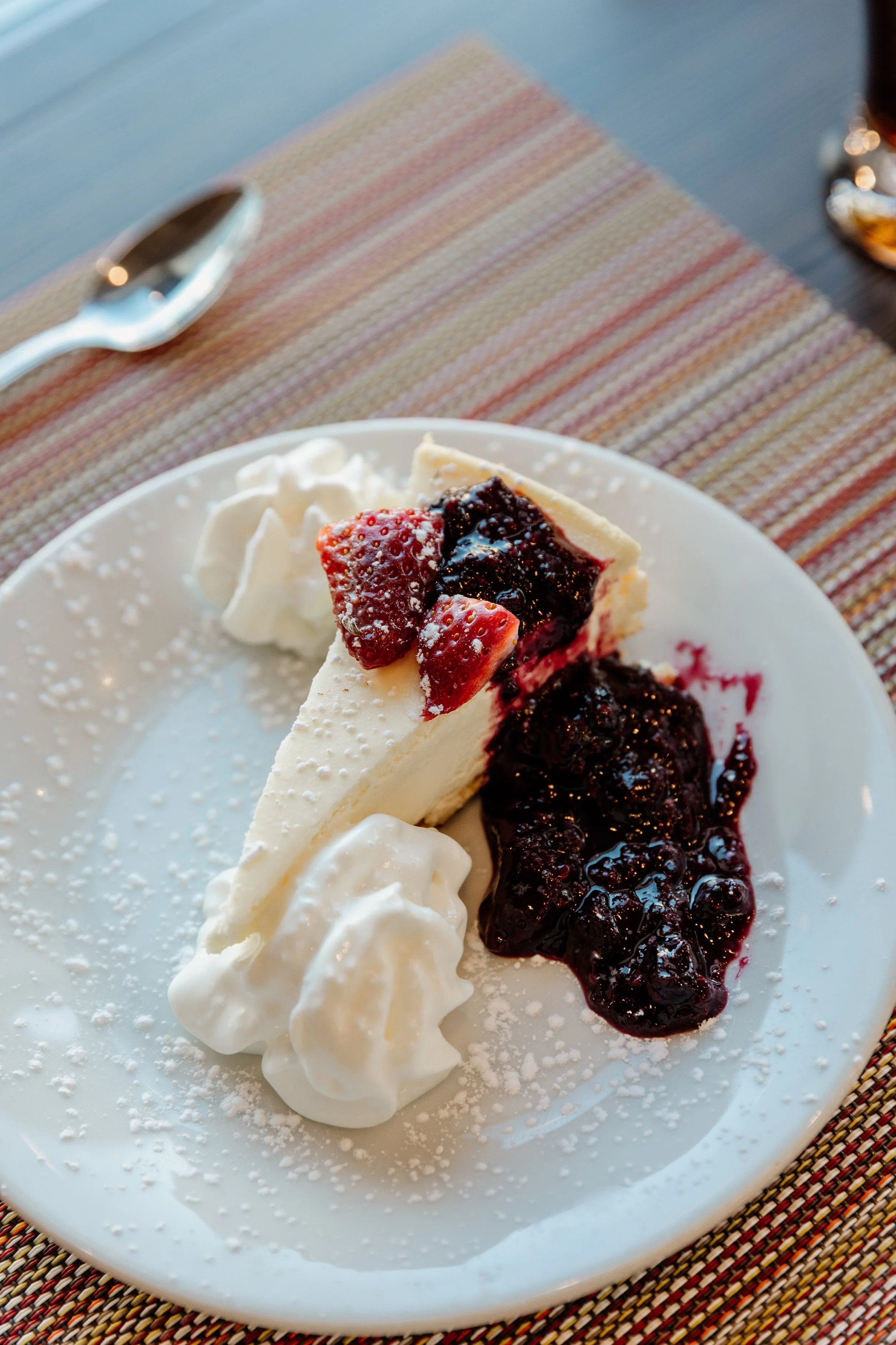 Slice of cheesecake with strawberry and blueberry topping, whipped cream, and powdered sugar on a white plate.