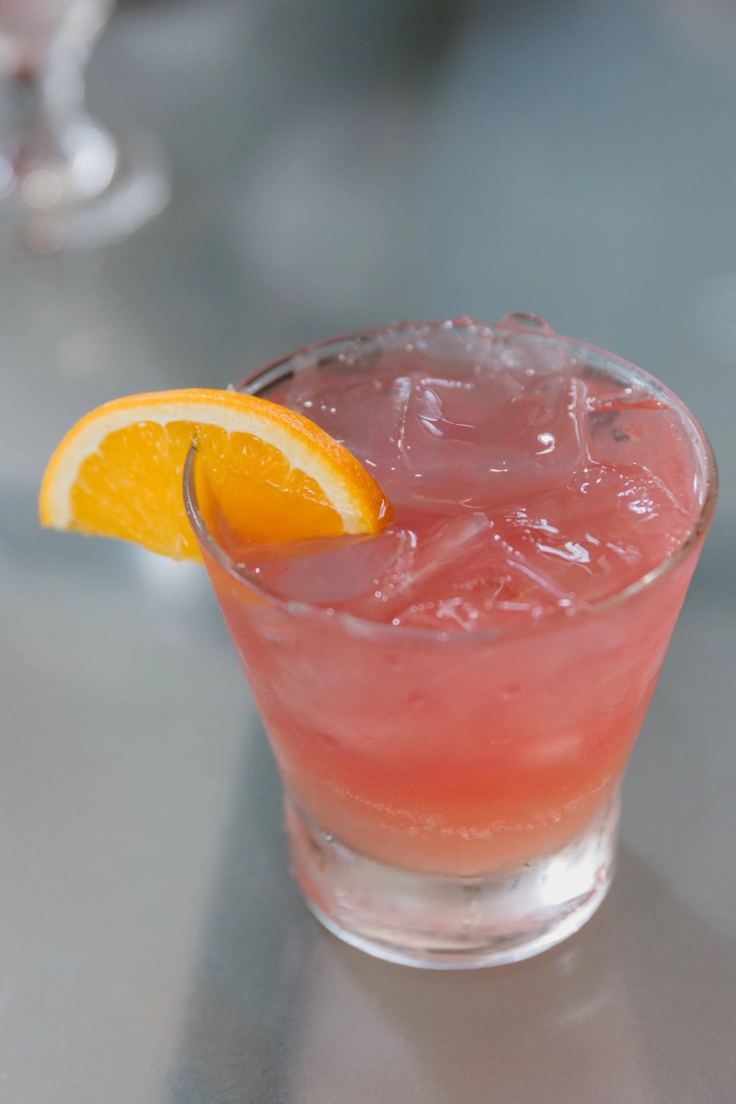 A glass of pink cocktail garnished with an orange wedge on a gray surface.