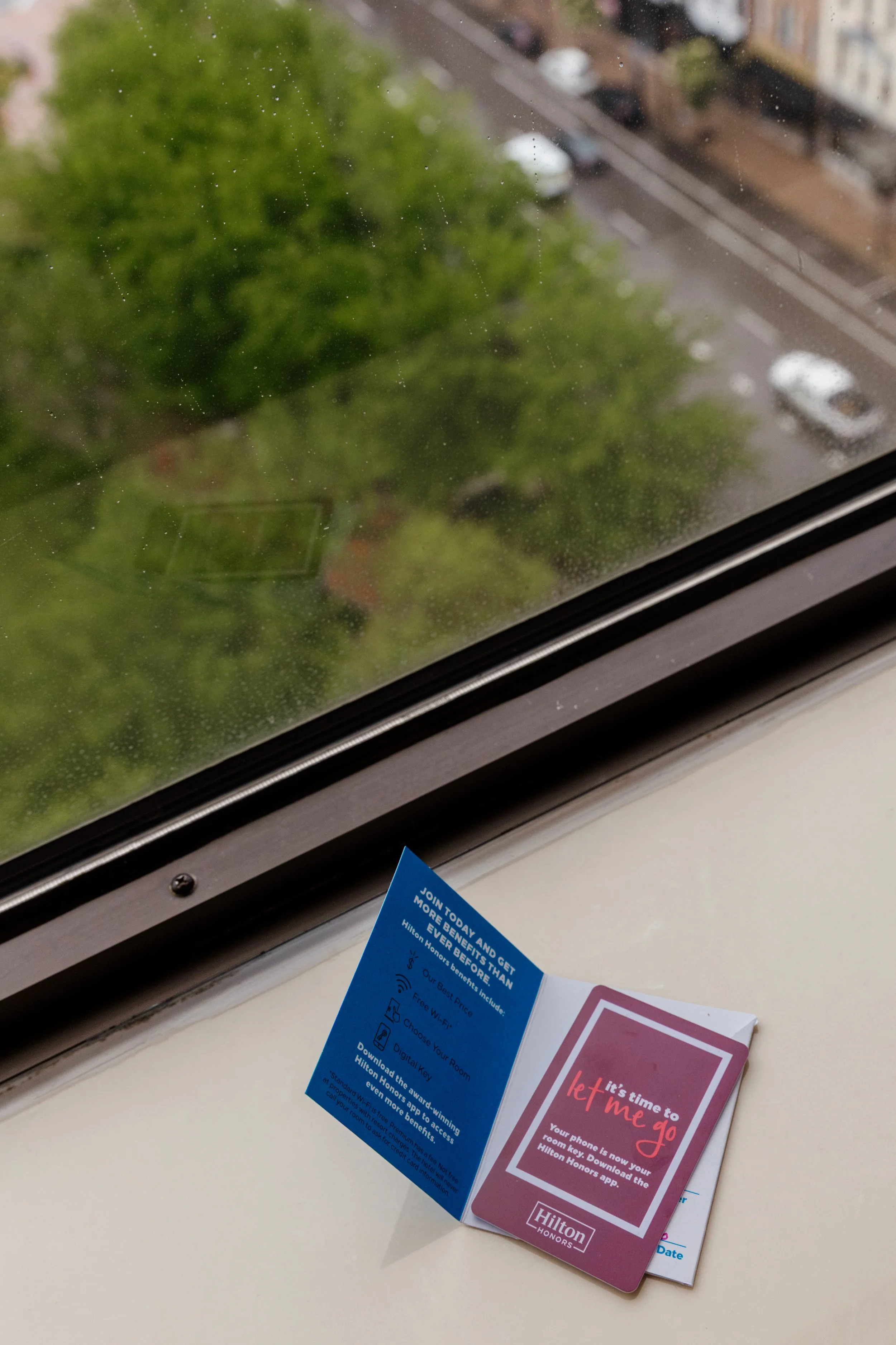 A window with a view of a tree and street below. On the window sill, there are Hilton Honors informational leaflets, including one with the words 'let me go' and a blue card with promotional text.