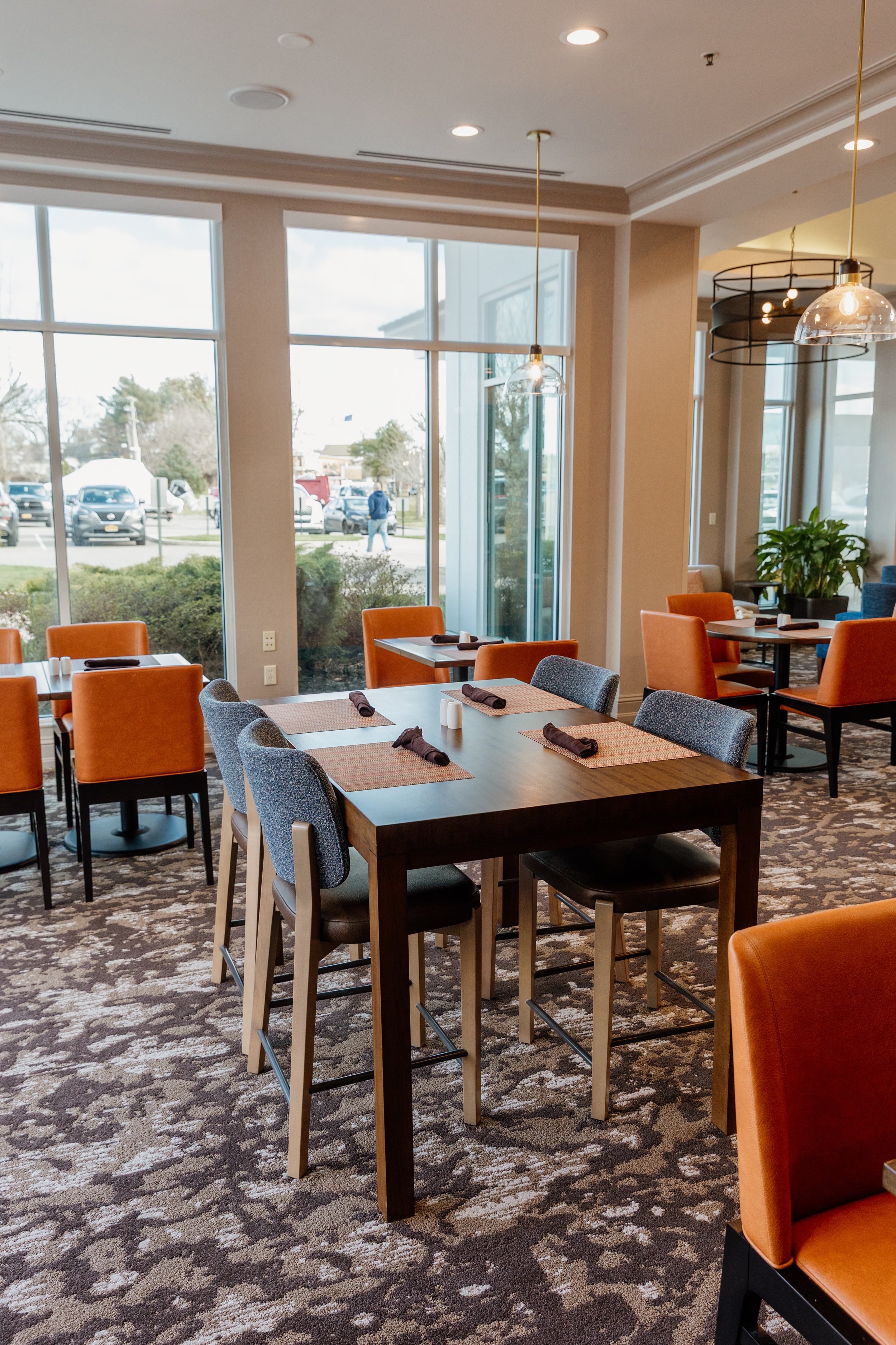 Interior of a restaurant dining area with tables set with placemats, rolled napkins, and salt and pepper shakers. Large windows bring in natural light, and there are orange and gray chairs. The floor has a patterned carpet, and there are hanging pend