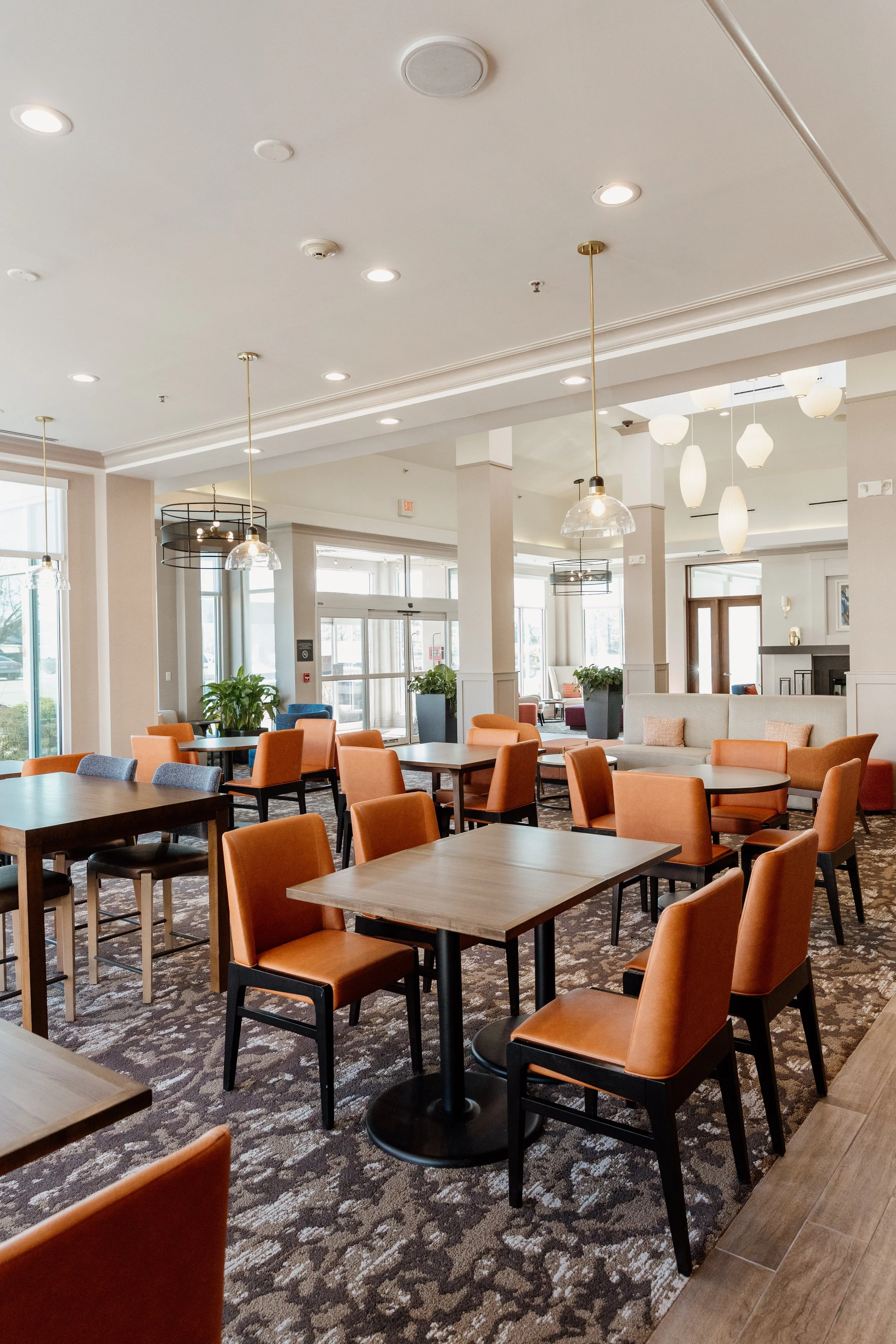 Interior of a spacious, well-lit dining or lounge area with multiple tables and orange chairs, decorative hanging lights, large windows, and potted plants.