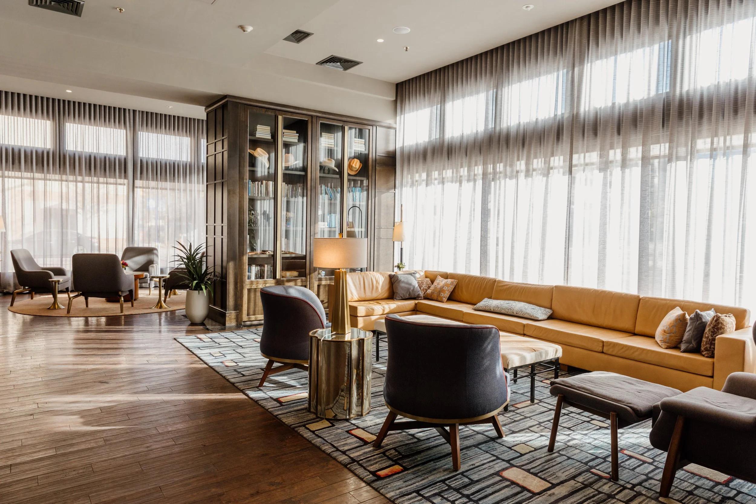 A modern hotel lounge with large windows, a yellow sectional sofa, black and gray armchairs, a wooden bookshelf with books and hats, and a patterned area rug.