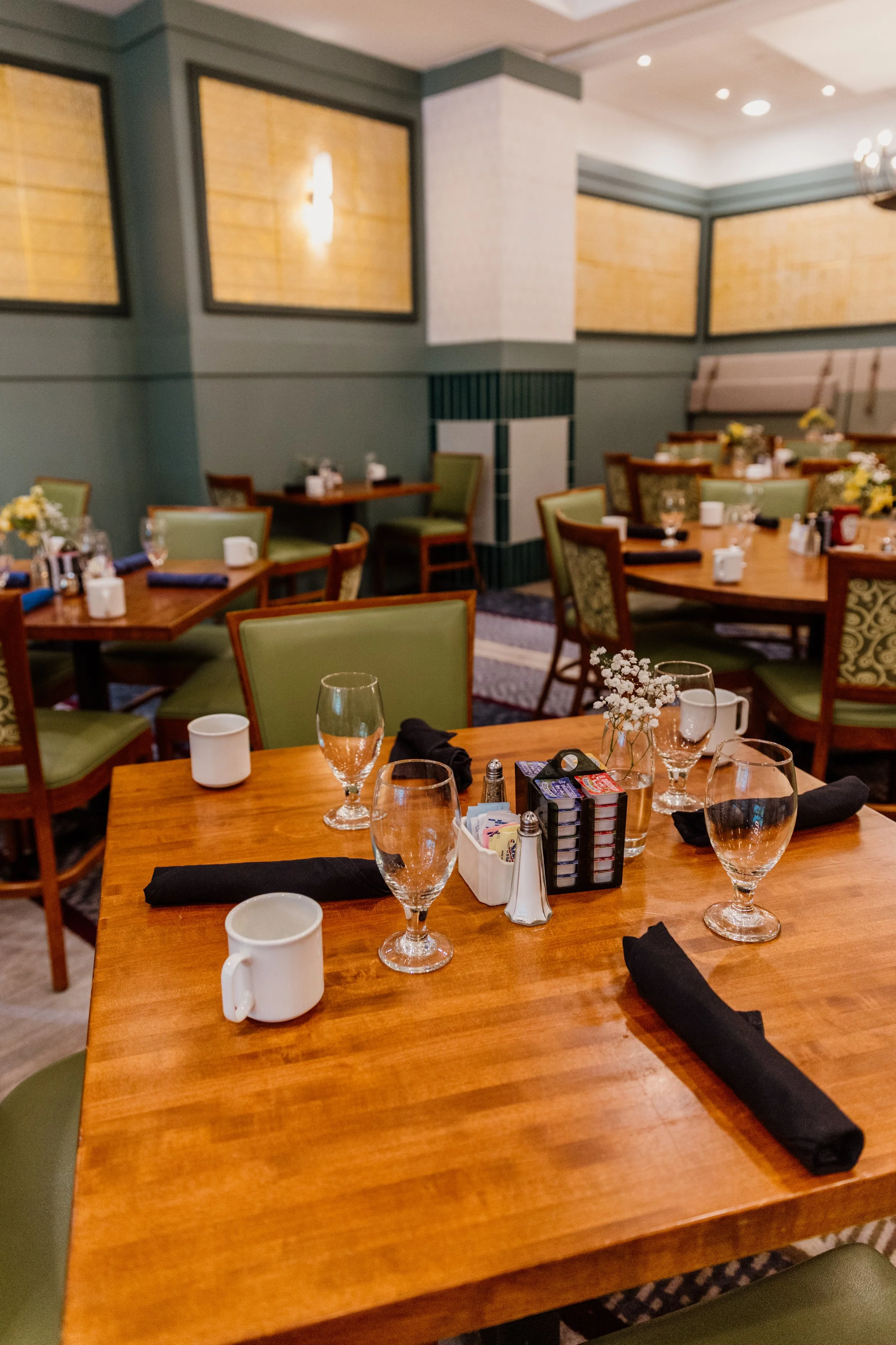 A dining room with multiple tables set with water glasses, coffee mugs, black cloth napkins, and small flower vases. The room has green chairs with patterned and plain upholstery and warm lighting.