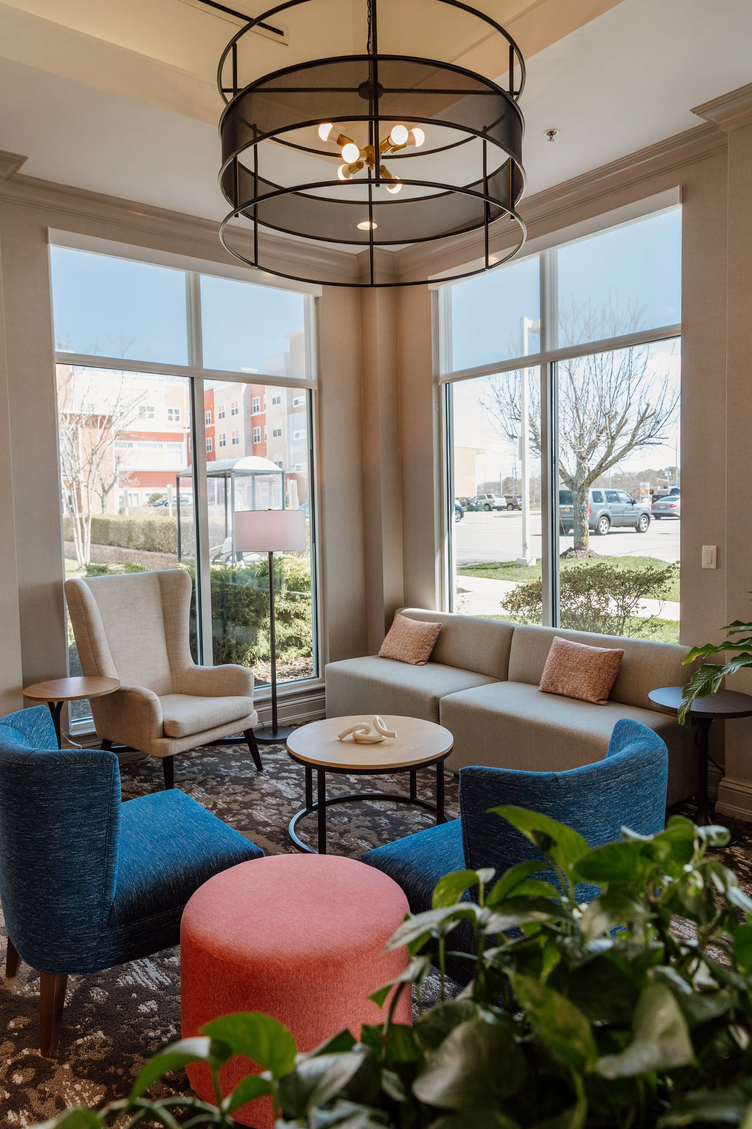Cozy sitting area with a white armchair, a beige sofa with pink cushions, blue armchairs, a pink ottoman, a small round coffee table with an abstract sculpture, a tall floor lamp, large windows with views of a parking lot and trees, a modern ceiling 