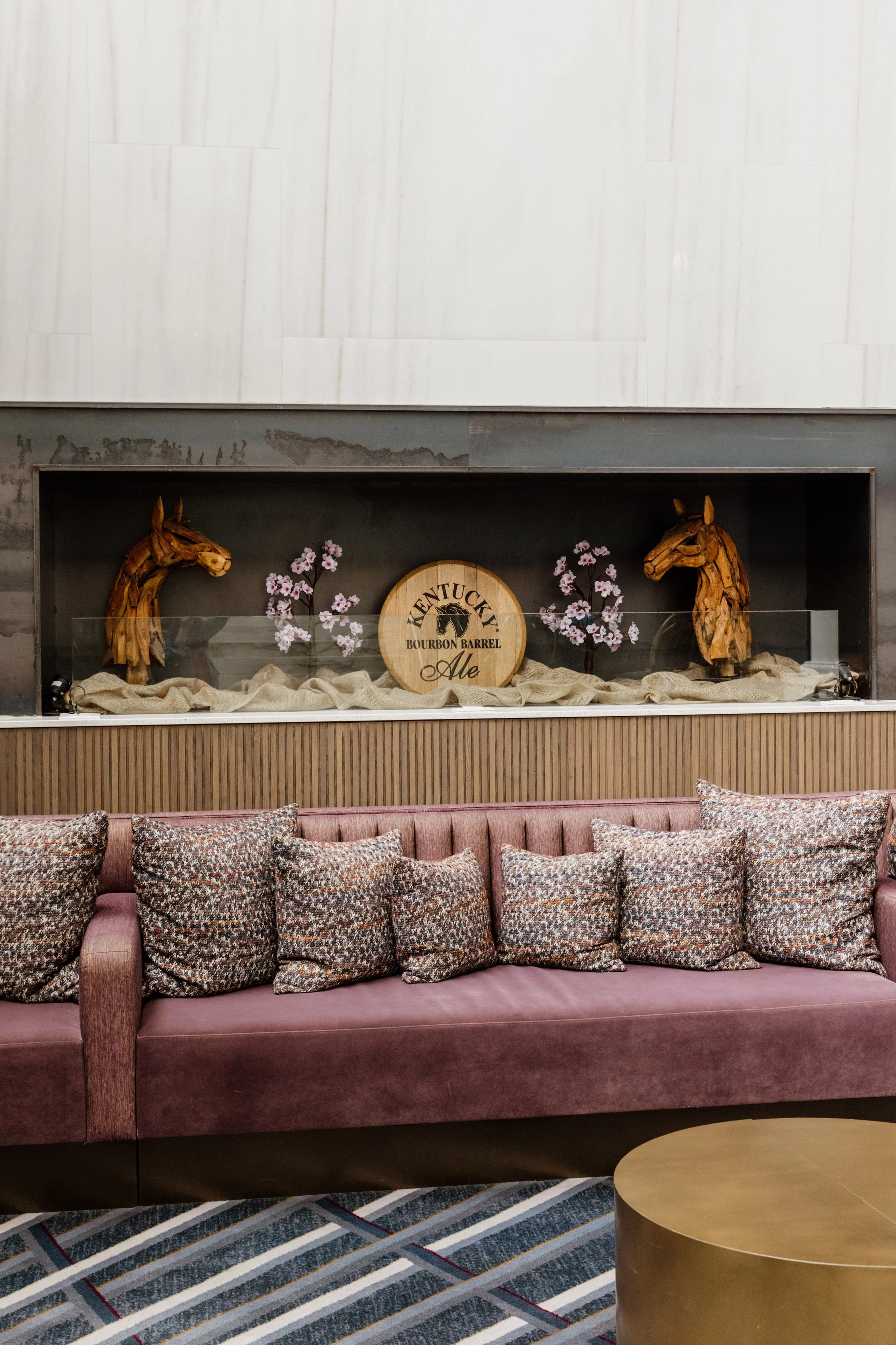 Decorative display with wooden horse heads, a Kentucky Bourbon Barrel Ale sign, pink cherry blossom branches, and beige fabric in a glass case above a mauve velvet sofa with patterned pillows, next to a round gold table.
