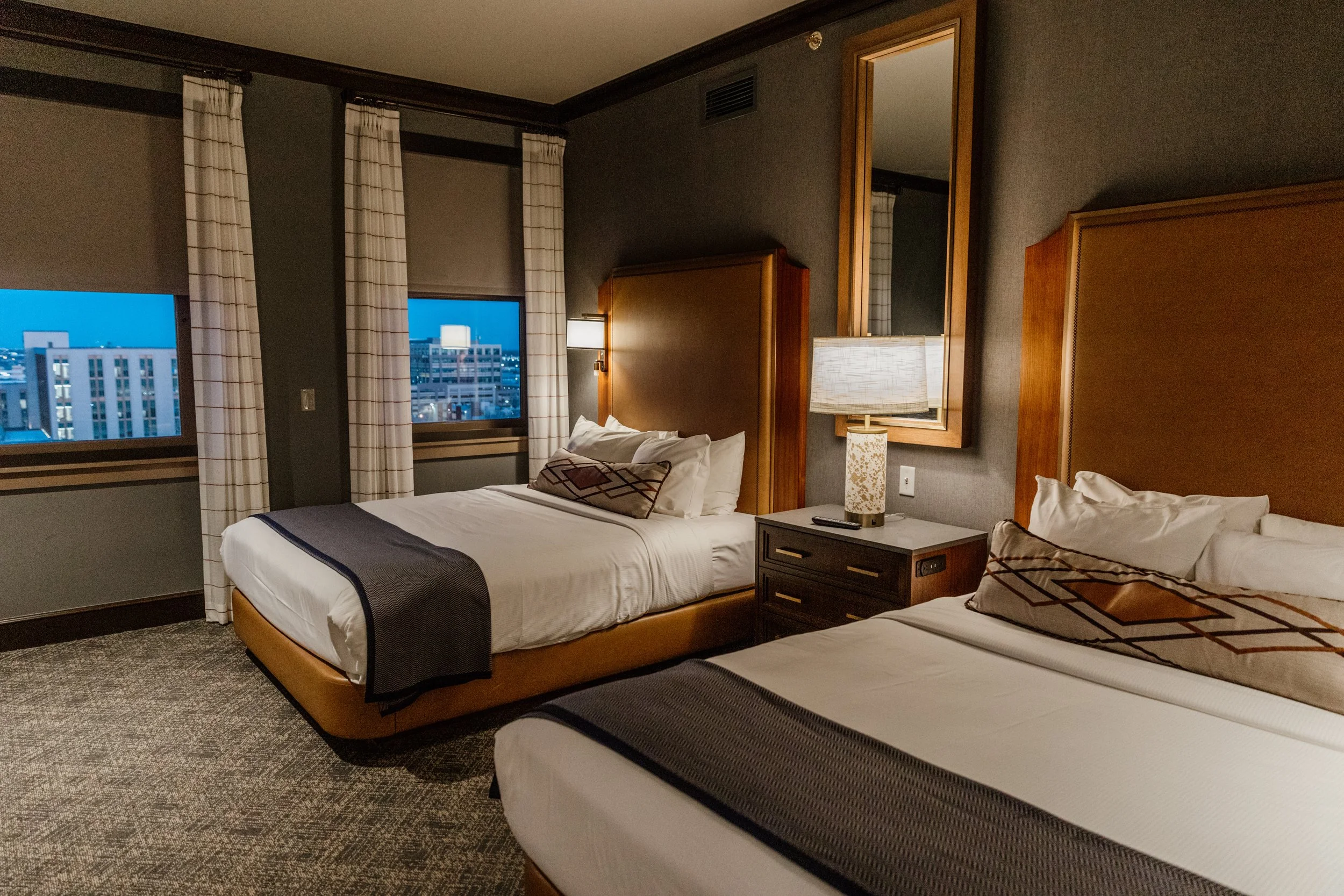 A hotel room with two beds, nightstand, lamp, and city view through windows.