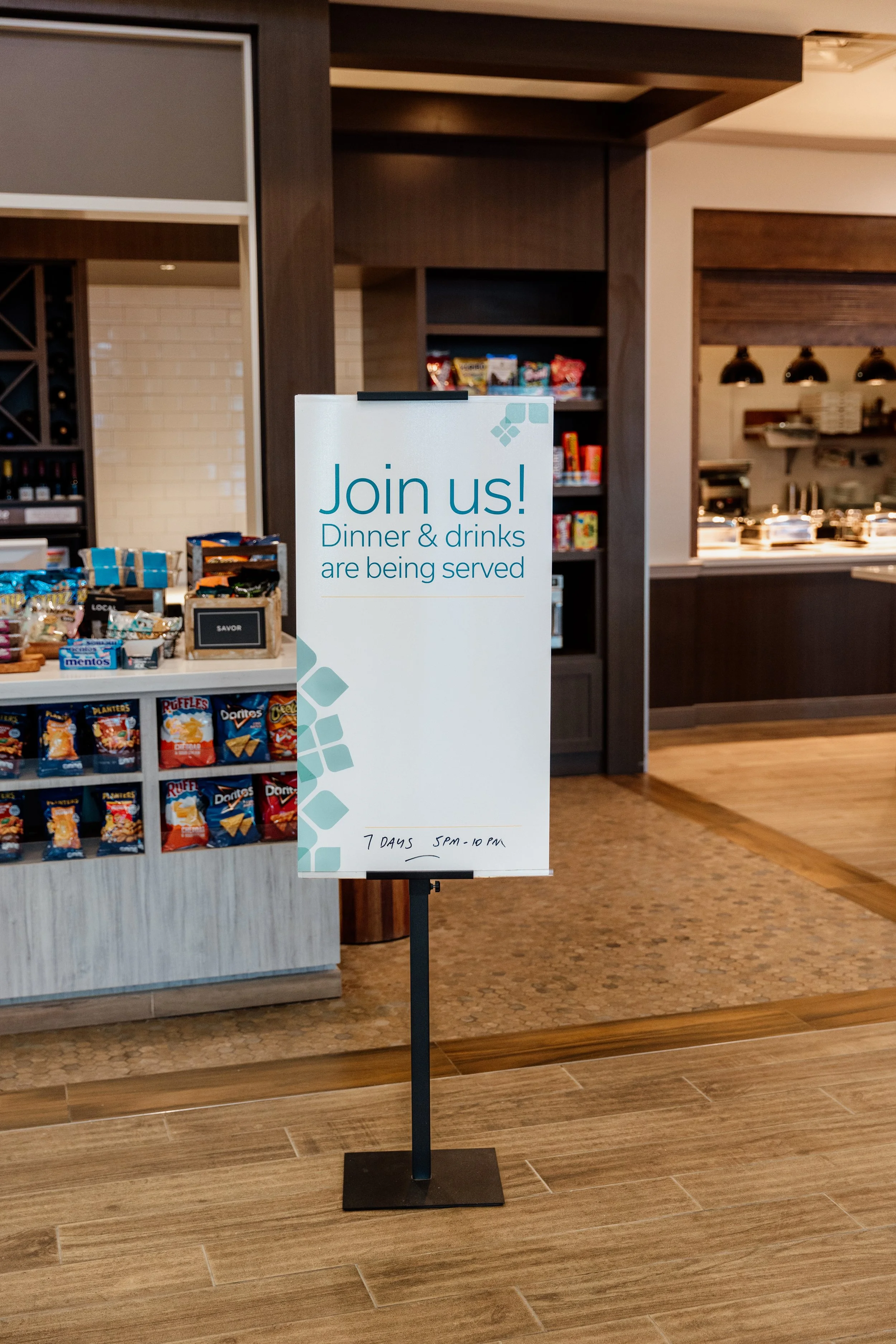 Restaurant sign inviting guests to join for dinner and drinks, with hours of 5 PM to 10 PM, located in a dining area with snack shelves and kitchen in the background.