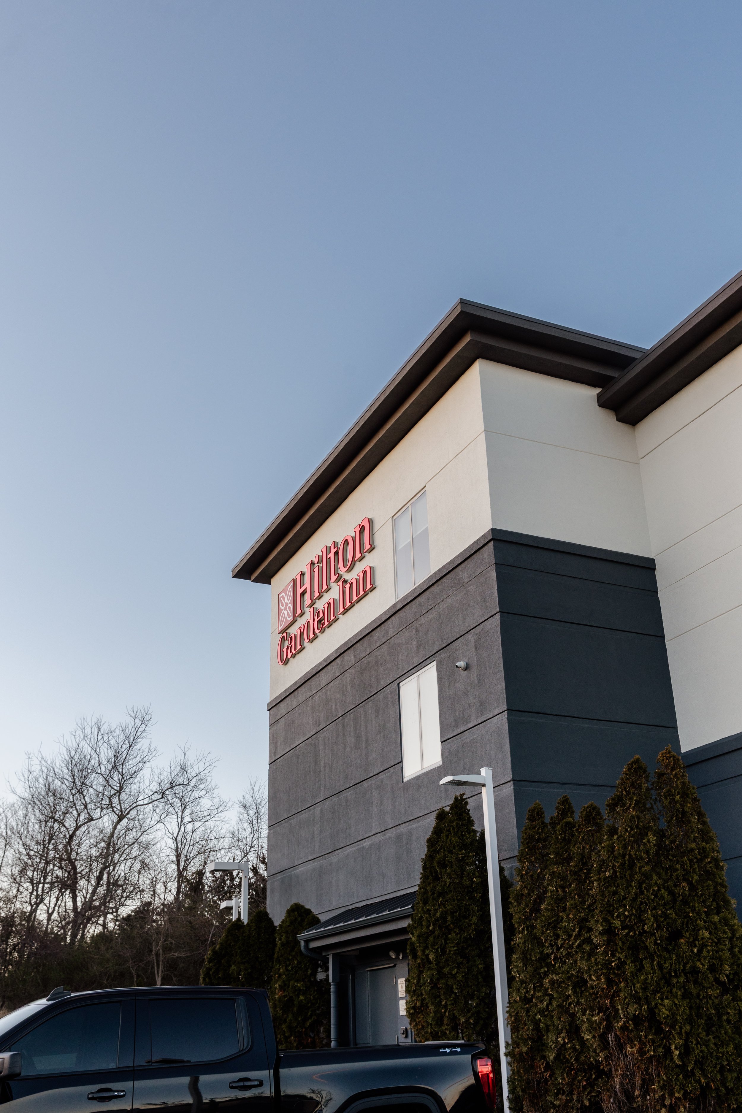 Exterior of a Hilton Garden Inn hotel building with a sign on the top corner, trees, a black pickup truck parked in front, and clear sky.