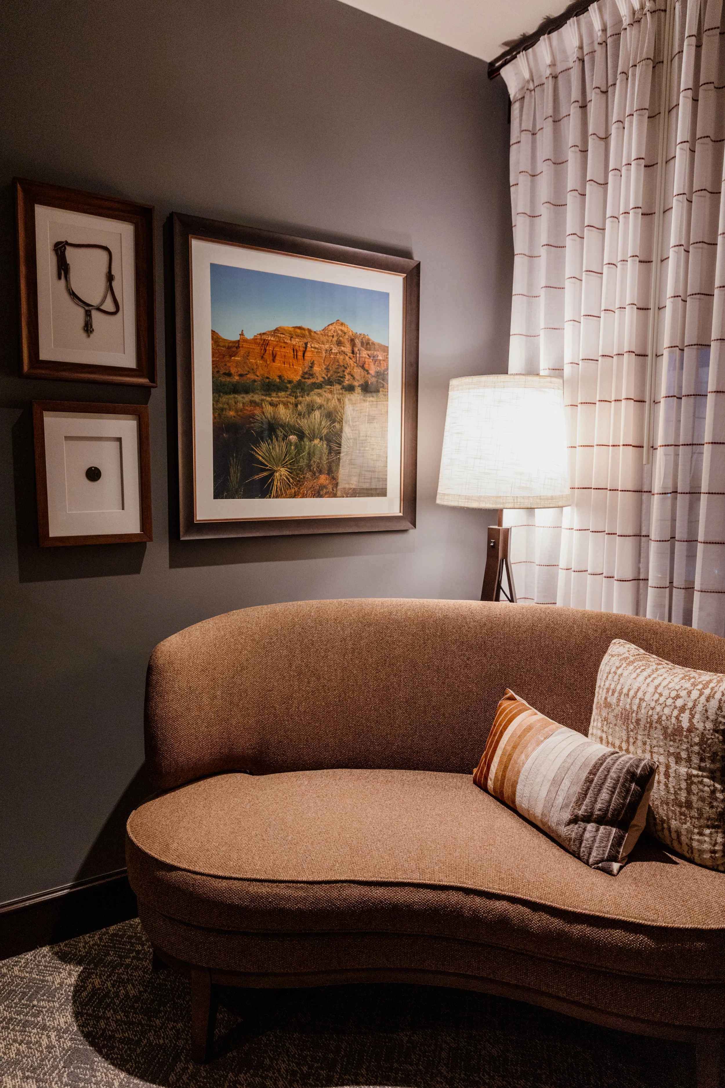 A cozy living room corner featuring a beige curved sofa with two decorative pillows, a tall floor lamp with a white lampshade, a large framed landscape photograph of a desert scene with red rock formations, and three smaller framed pictures on a gray