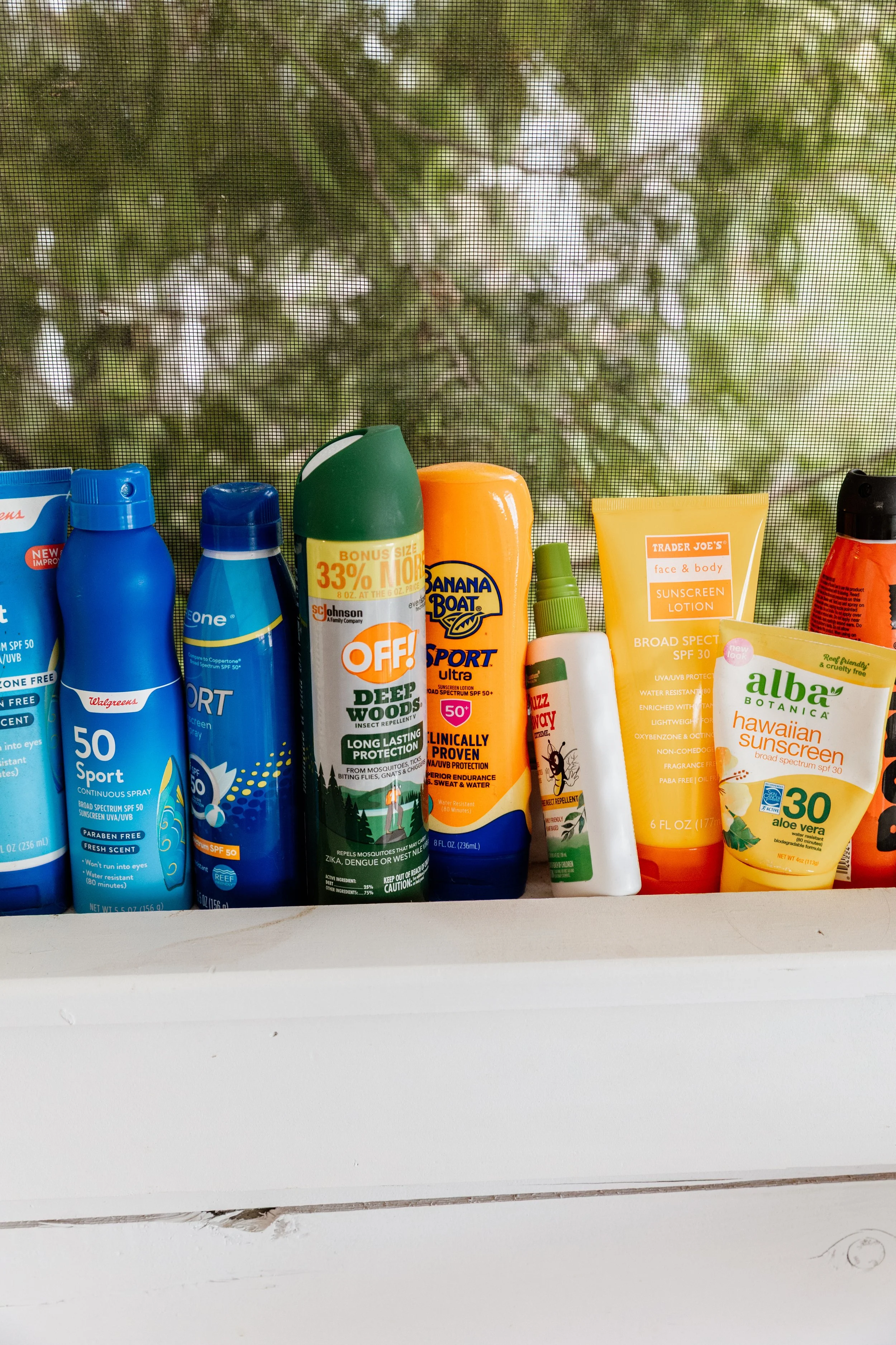 Assorted bottles and tubes of sunscreen and insect repellent on a white surface, with a screened window and green foliage outside