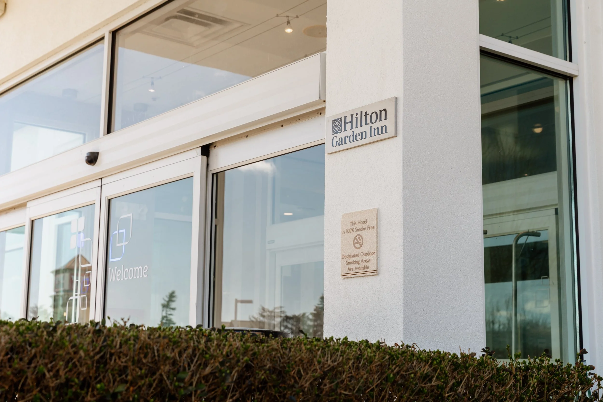 Exterior of Hilton Garden Inn hotel with white walls, glass doors, and signs indicating the hotel name and smoke-free policy.