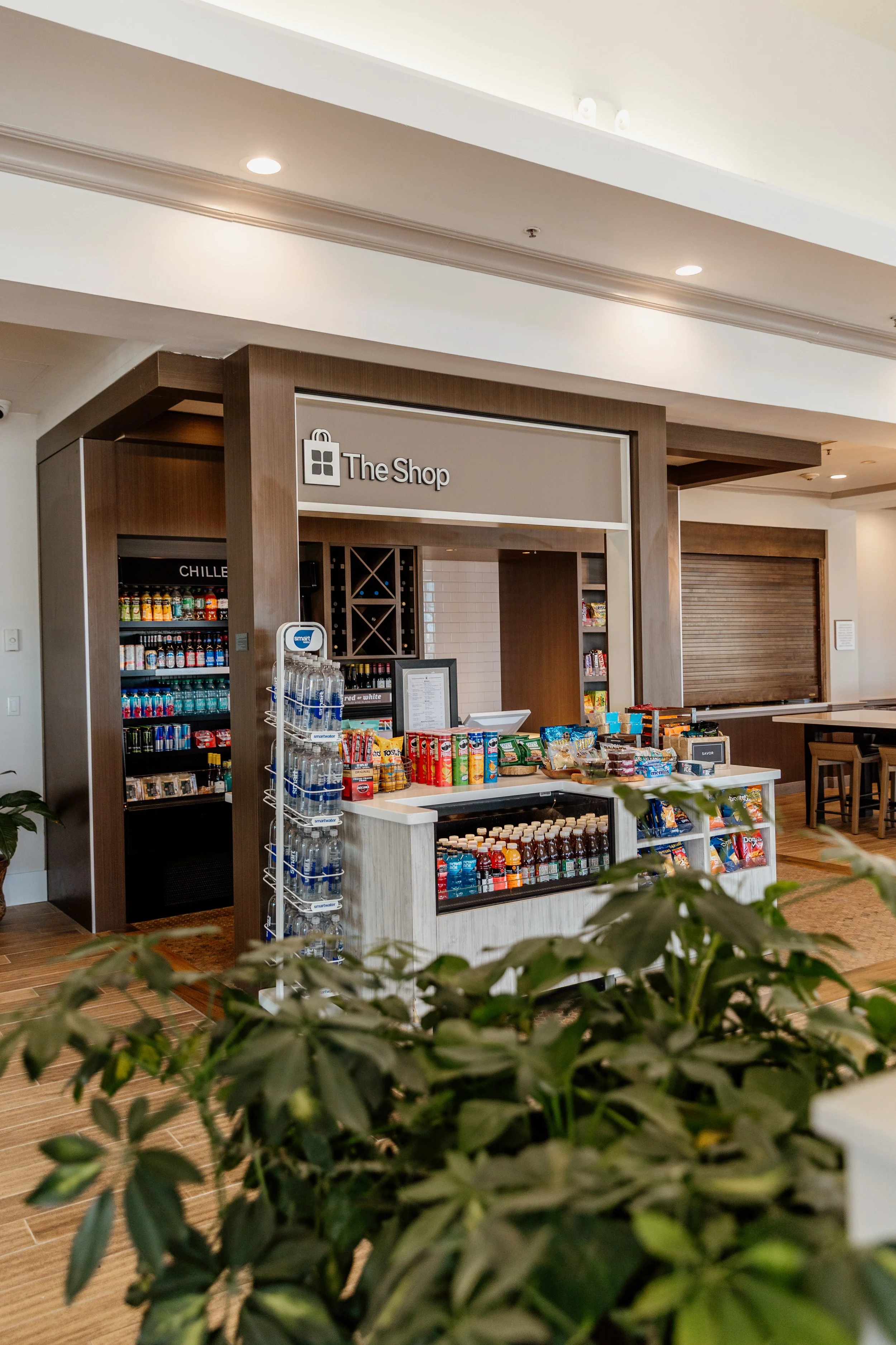 A small retail shop called "The Shop" inside a building, featuring shelves stocked with snacks, drinks, and bottled beverages, with a countertop display and a water bottle stand in front.