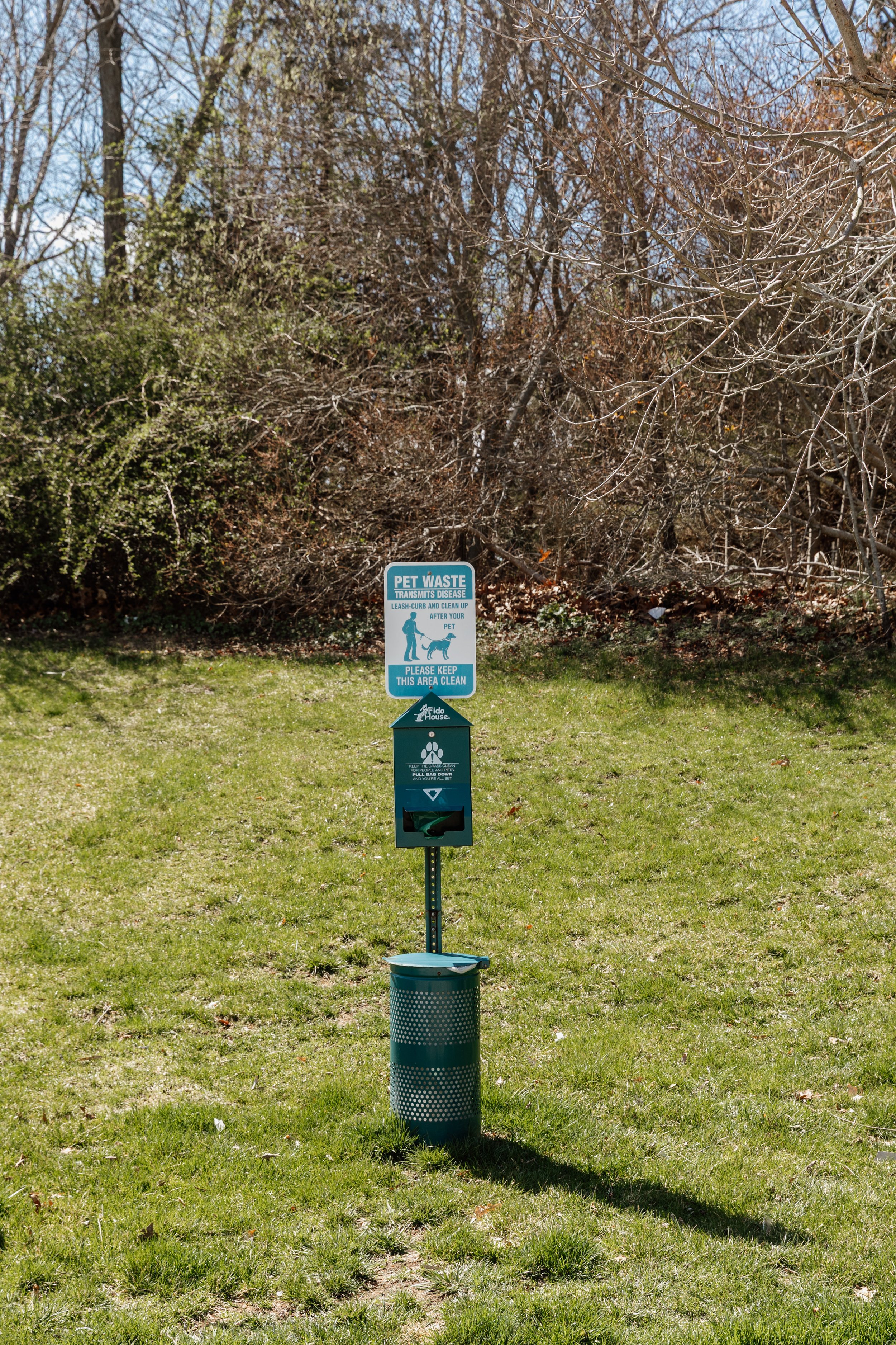 Dog waste station with a sign that reads 'Pet Waste Transmits Disease' and instructions to leash, curb, and clean up after your pet, with a waste bag dispenser and a trash can nearby, on a grassy area with trees in the background.