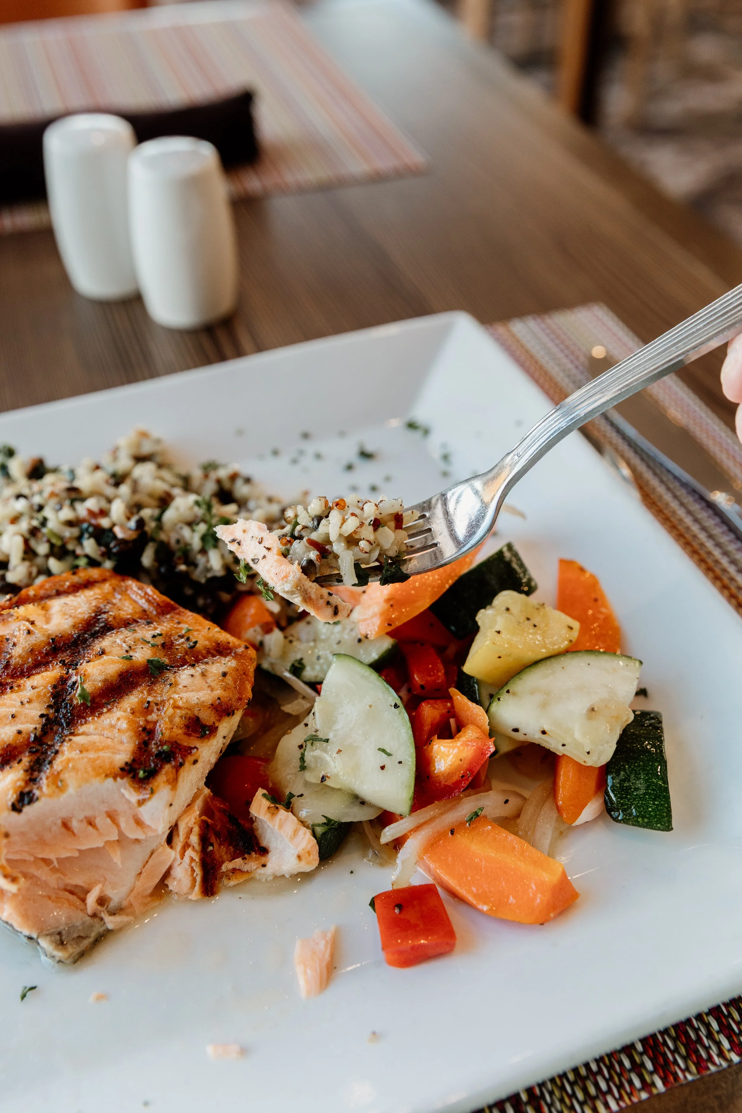 Close-up of a plate of grilled salmon with roasted vegetables and a side of quinoa salad on a white square plate, with salt and pepper shakers in the background.